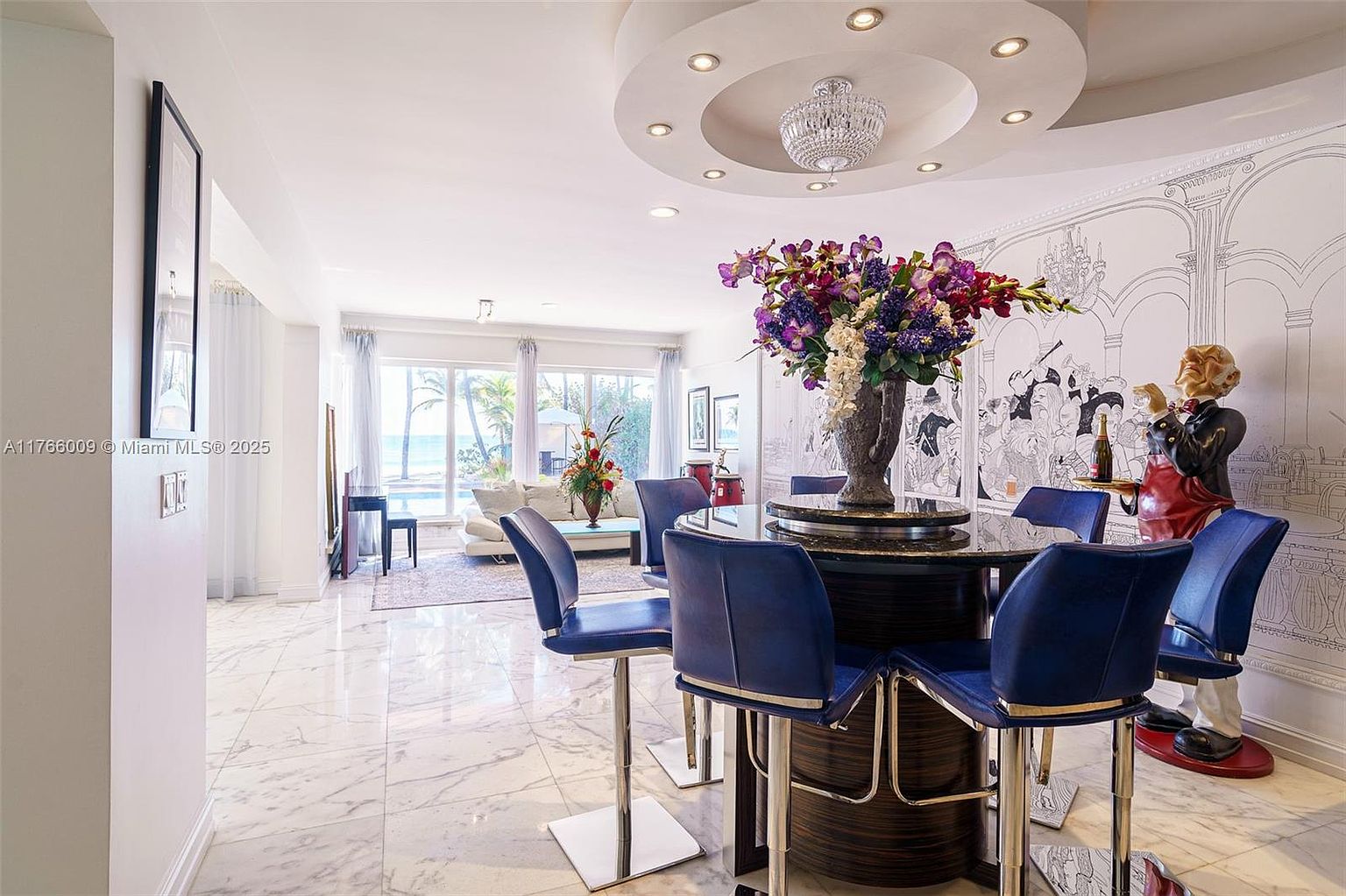 This elegant dining area features a circular table surrounded by four vibrant blue chairs, set against a backdrop of a whimsical, hand-drawn style mural. A large, ornate crystal chandelier hangs from a recessed circular ceiling feature, casting a sophisticated glow over the space. The room flows seamlessly into a bright living area with large windows that provide a view of the outdoor greenery and pool area.