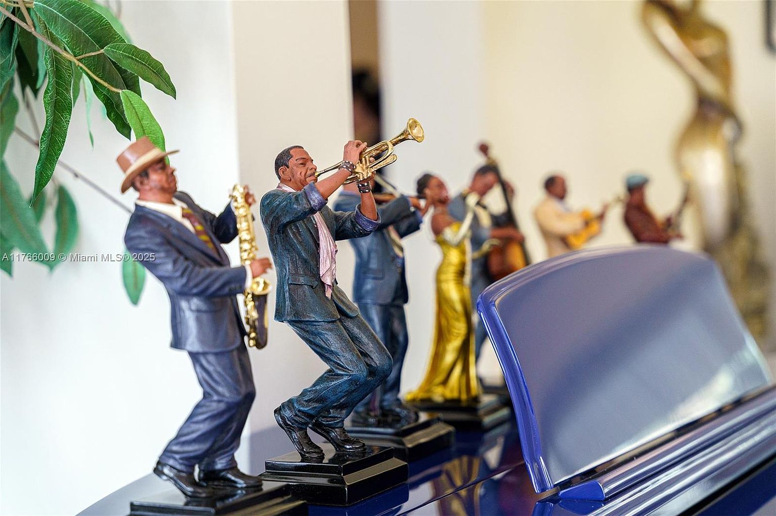 This close-up shot features a collection of detailed jazz band figurines arranged on a polished, dark blue surface, likely the lid of a grand piano. The figures include musicians playing a saxophone, trumpet, and double bass, rendered with expressive poses and formal attire. The shallow depth of field keeps the focus on the musicians while softly blurring the background, which includes a glimpse of a decorative sculpture.