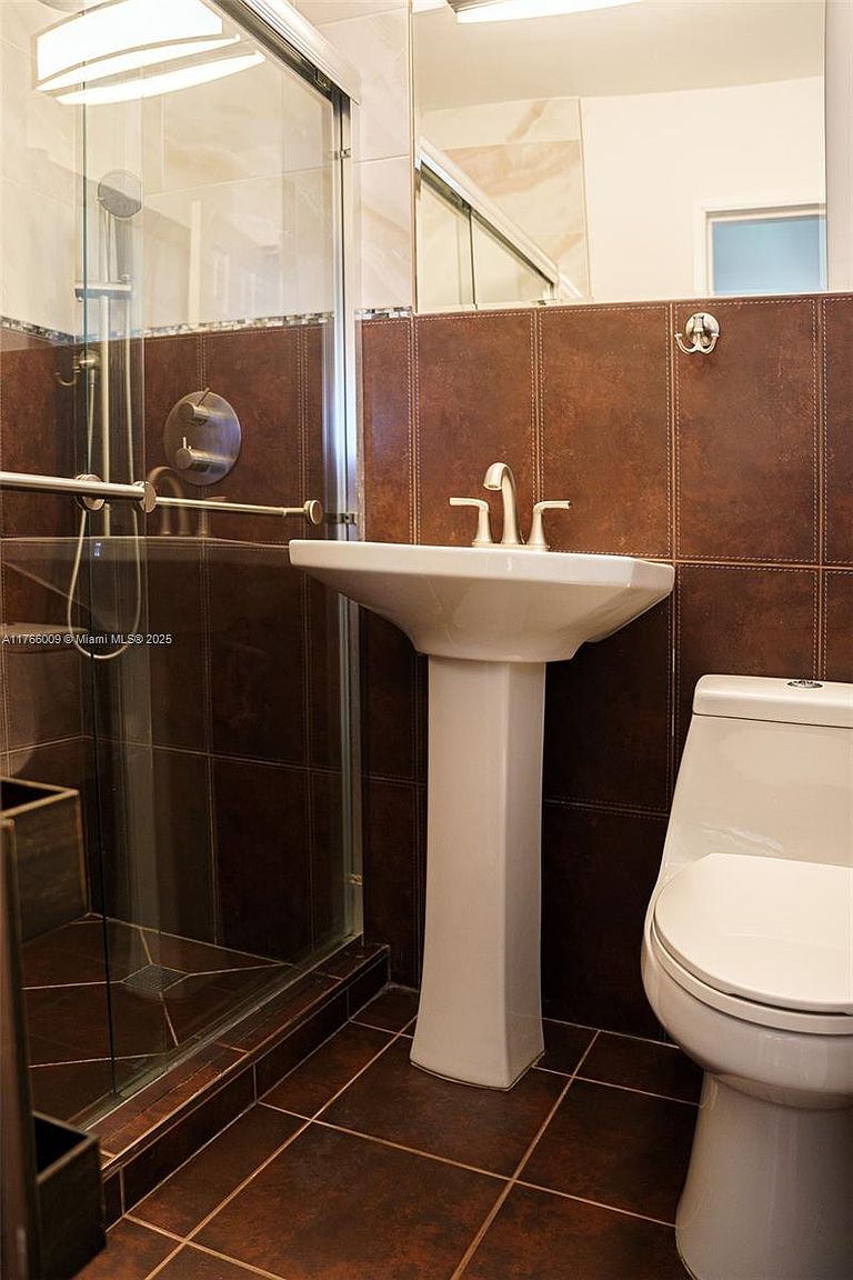 This compact bathroom features dark, earth-toned wall and floor tiles that create a moody, cohesive aesthetic. The space includes a white pedestal sink with a chrome faucet, a modern toilet, and a glass-enclosed shower stall. A large mirror above the sink reflects the room, enhancing the sense of space in this functional, contemporary bathroom.