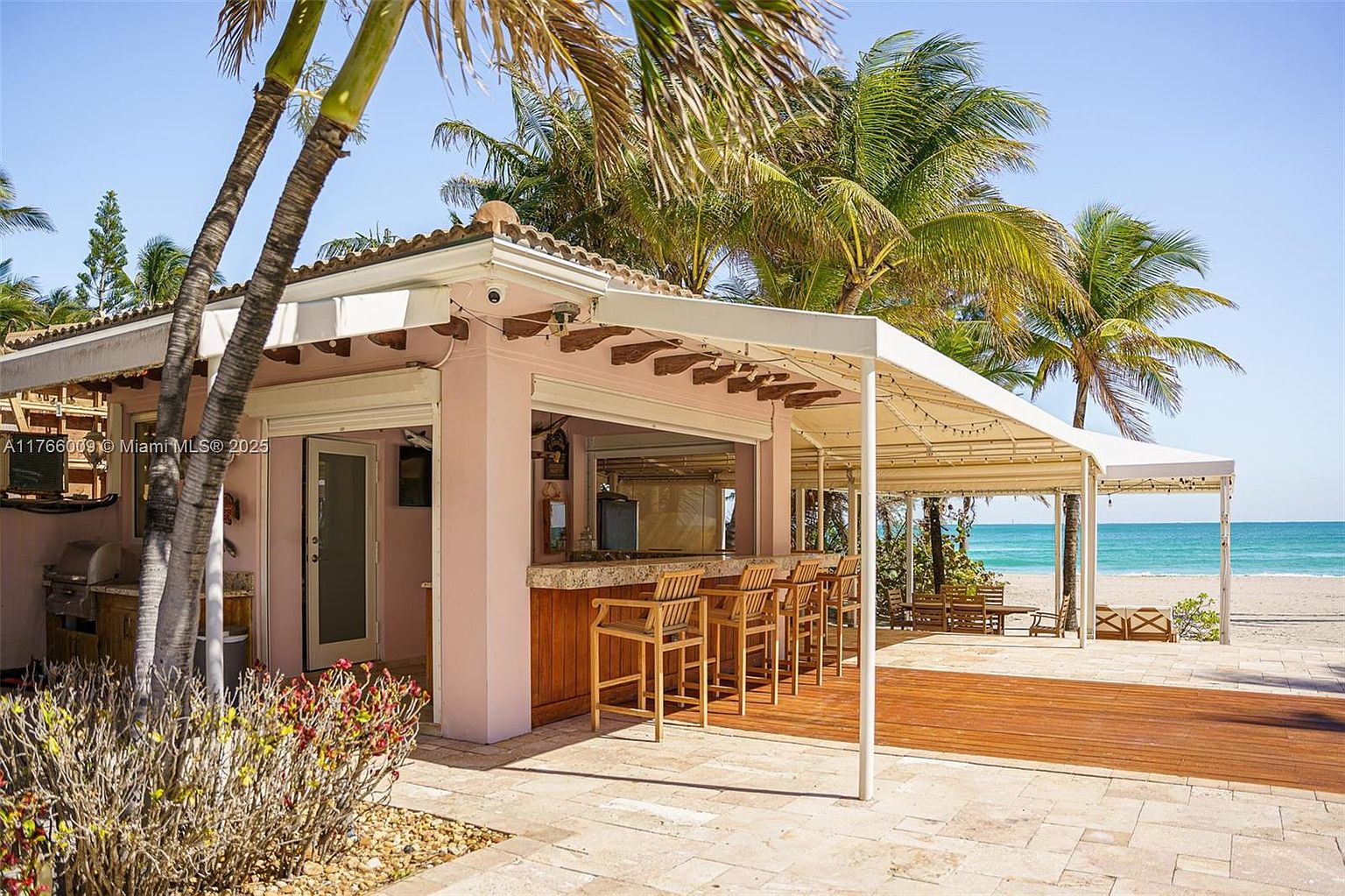 This outdoor living space features a covered bar area with wooden stools, set against a light-colored structure with a pergola-style roof. The patio transitions from stone pavers to a wooden deck, leading directly toward a sandy beach and the ocean. Lush palm trees frame the scene, creating a tropical, resort-like atmosphere perfect for entertaining.