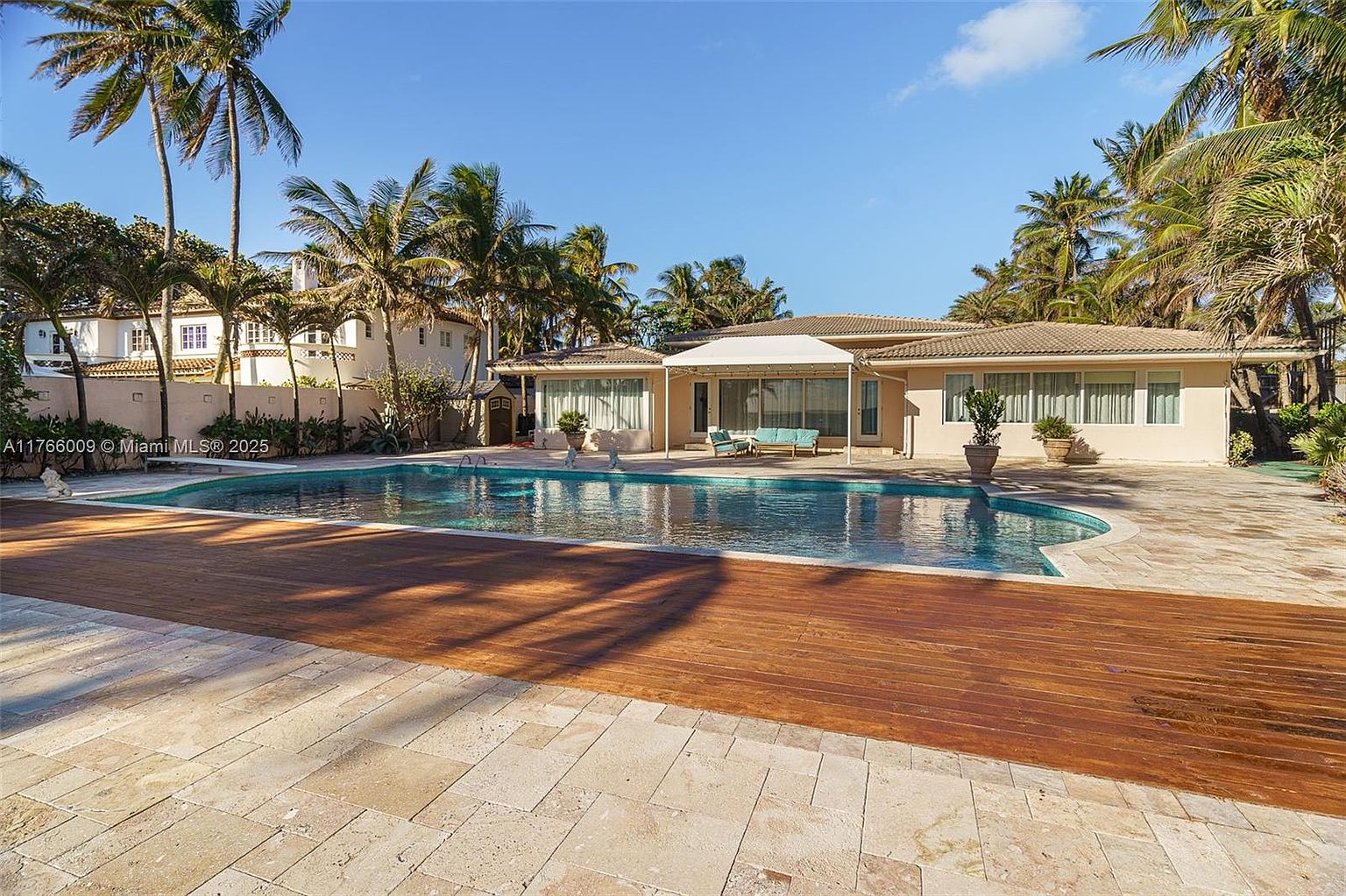 This expansive backyard features a large, free-form swimming pool as the central focal point, set against a backdrop of lush palm trees and a clear blue sky. The residence showcases a light-colored exterior with a covered patio area, comfortable outdoor seating, and a mix of stone and wood decking. The overall impression is one of a serene, tropical-inspired private oasis perfect for outdoor entertaining.