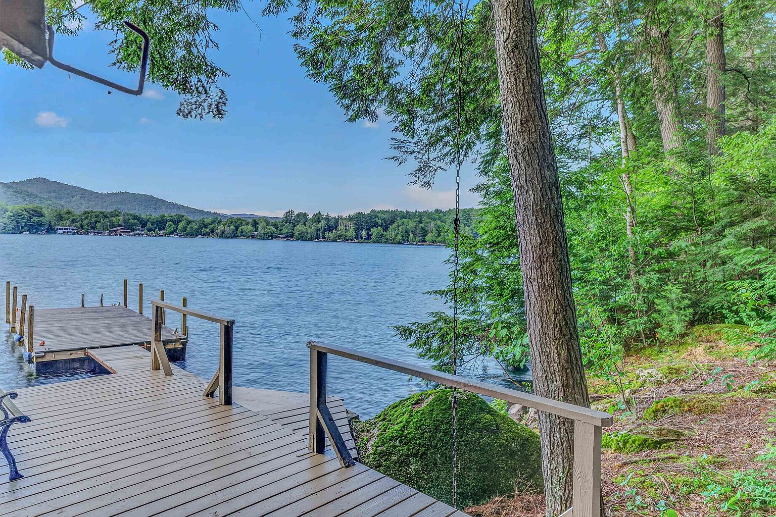 This image captures a serene wooden deck extending out over a calm lake, framed by lush green trees and a large moss-covered boulder. The perspective is from the deck looking out toward the water, showcasing a peaceful waterfront setting with distant mountains under a clear blue sky. The scene emphasizes a private, natural retreat perfect for relaxation or water-based activities.