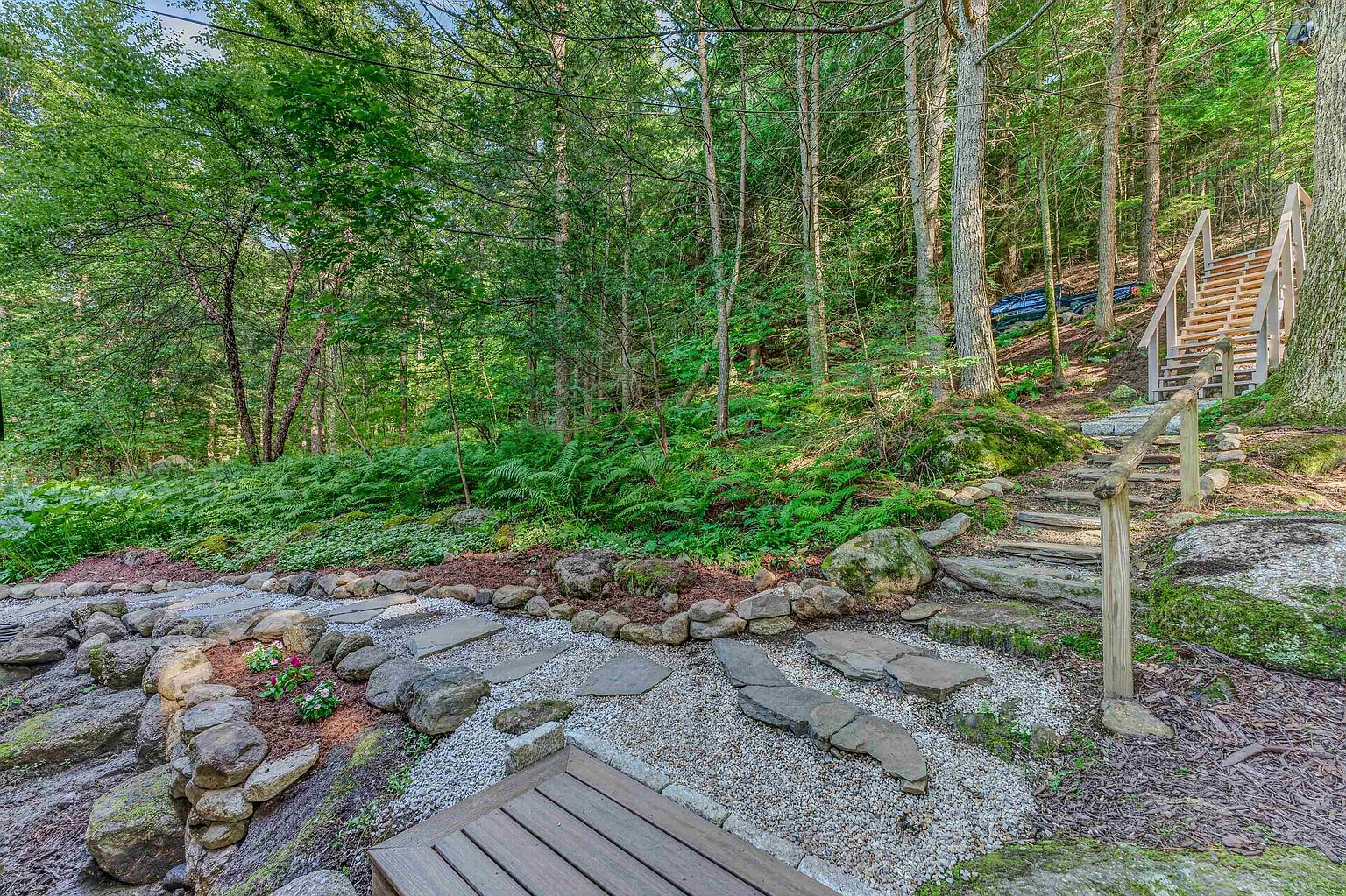 This serene outdoor space features a landscaped garden path with stone pavers set into gravel, leading toward a wooden staircase that ascends into a lush, wooded hillside. The area is surrounded by dense forest greenery, ferns, and natural rock formations, creating a peaceful and secluded atmosphere. The perspective is from a low angle near a wooden deck, emphasizing the transition from the manicured garden to the natural woodland setting.