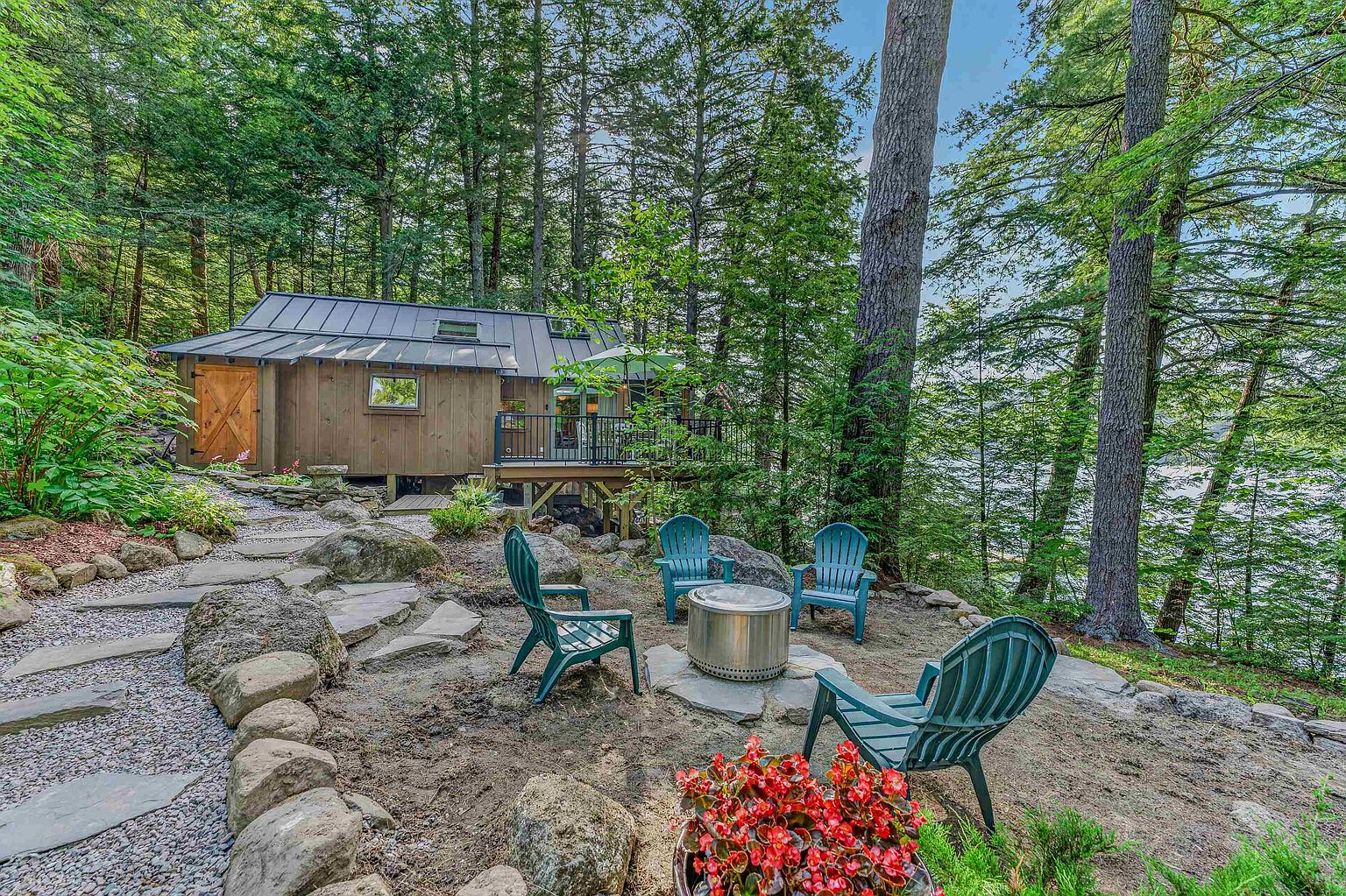 This serene outdoor space features a rustic cabin nestled among tall trees, overlooking a body of water. In the foreground, a stone-paved seating area includes four Adirondack chairs arranged around a modern metal fire pit, accented by a vibrant planter of red flowers. The scene captures a peaceful, wooded retreat with a natural landscape design that seamlessly integrates the home with its lakeside environment.
