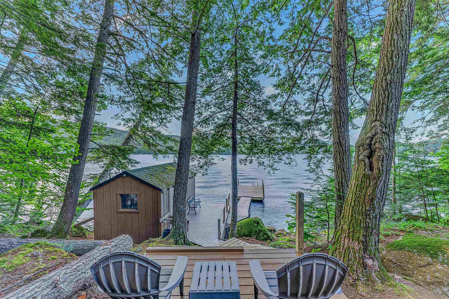 This scenic outdoor space features a wooden deck in the foreground with two dark chairs, overlooking a tranquil lake surrounded by lush, mature trees. A small, rustic wooden cabin sits near the water's edge, connected to a private dock that extends into the calm lake. The perspective is captured from an elevated vantage point, emphasizing the peaceful, secluded nature of this waterfront property.