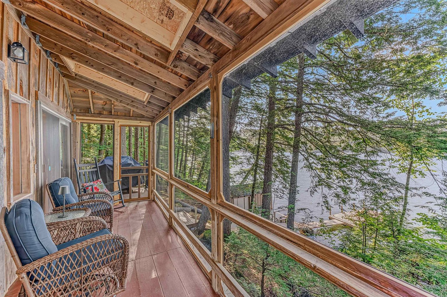 This screened-in porch offers a serene, rustic retreat with a direct view of a tranquil lake surrounded by dense forest. The space is furnished with comfortable wicker chairs and a small side table, creating an inviting spot for relaxation. The wooden construction and mesh screening provide an immersive connection to the natural environment, emphasizing the peaceful, secluded atmosphere of the property.