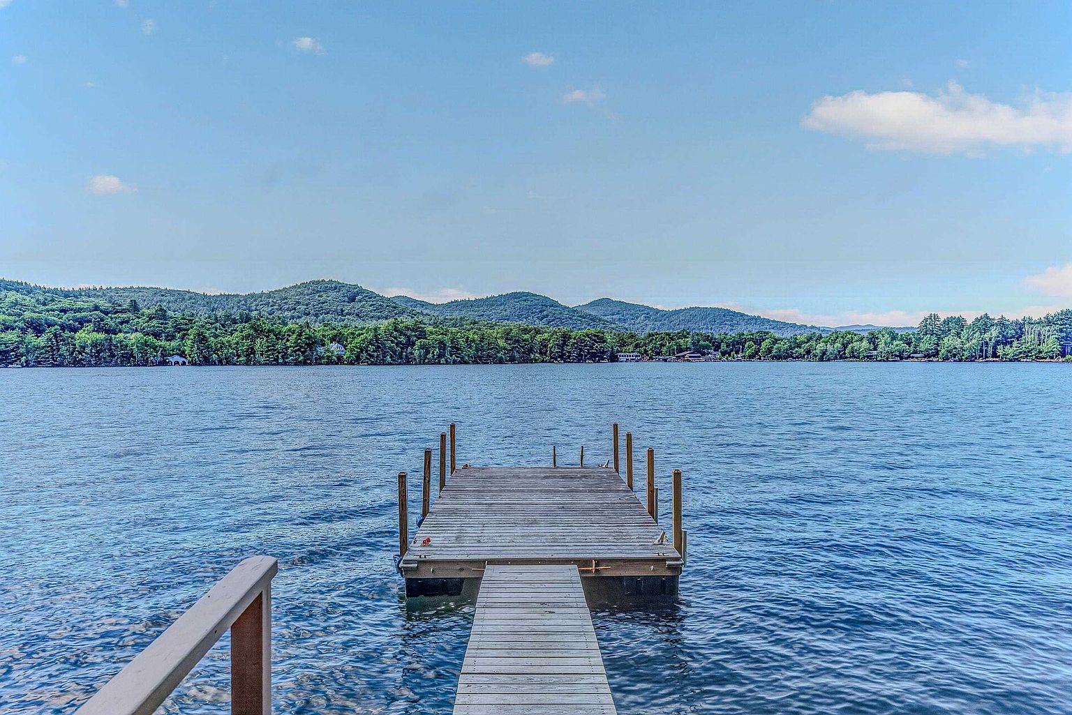This image captures a serene, eye-level perspective of a wooden dock extending into a calm, expansive lake. In the background, lush green mountains rise under a bright blue sky with scattered clouds, creating a peaceful and picturesque waterfront setting. The scene emphasizes the property's direct access to nature and recreational water activities.