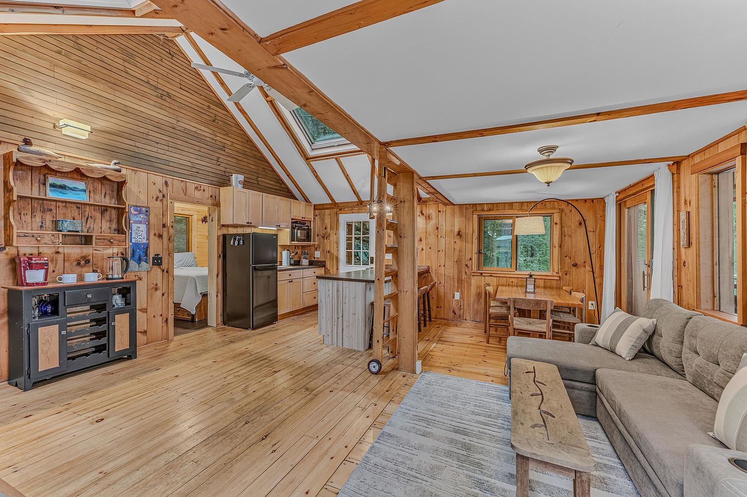 This open-concept living space features warm wood-paneled walls and ceilings, creating a cozy, rustic cabin atmosphere. The room includes a comfortable gray sectional sofa, a wooden coffee table, and a dining area, all illuminated by a combination of natural light from a skylight and warm interior lighting. A kitchen with a black refrigerator and light wood cabinetry is visible in the background, seamlessly integrated into the main living area.