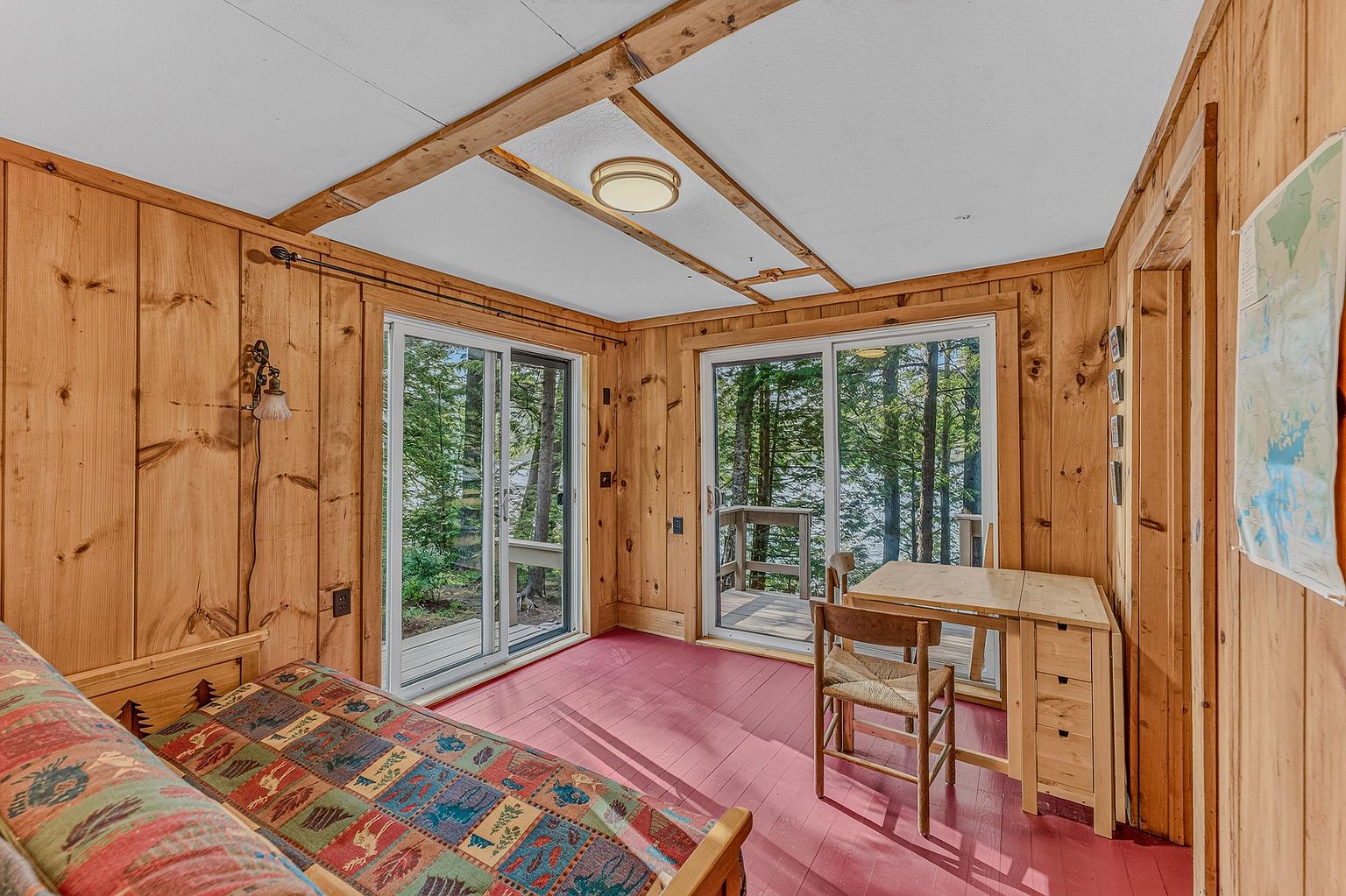 This rustic bedroom features warm wood-paneled walls and a vibrant, patterned quilt on the bed. Two sets of sliding glass doors provide abundant natural light and direct views of the surrounding forest and lake. A simple wooden desk and chair are positioned near the glass, creating a serene workspace within this cozy, cabin-style retreat.