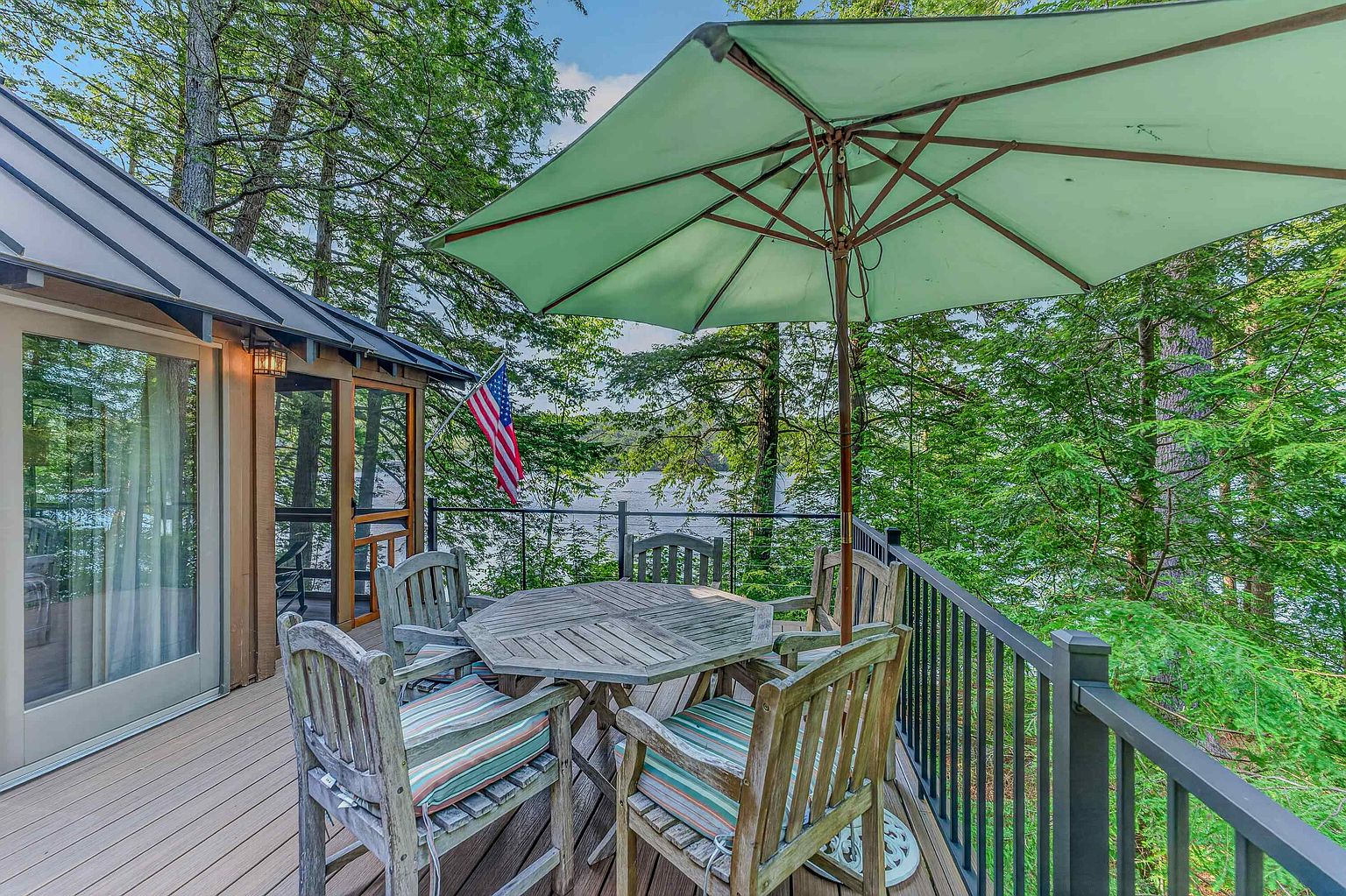 This elevated wooden deck offers a serene outdoor dining space overlooking a lush, tree-lined lake. The area is furnished with a hexagonal wooden table and matching chairs, shaded by a large light green umbrella, while a screened-in porch and sliding glass doors provide seamless indoor-outdoor access. An American flag is mounted to the railing, enhancing the peaceful, classic lakeside atmosphere.