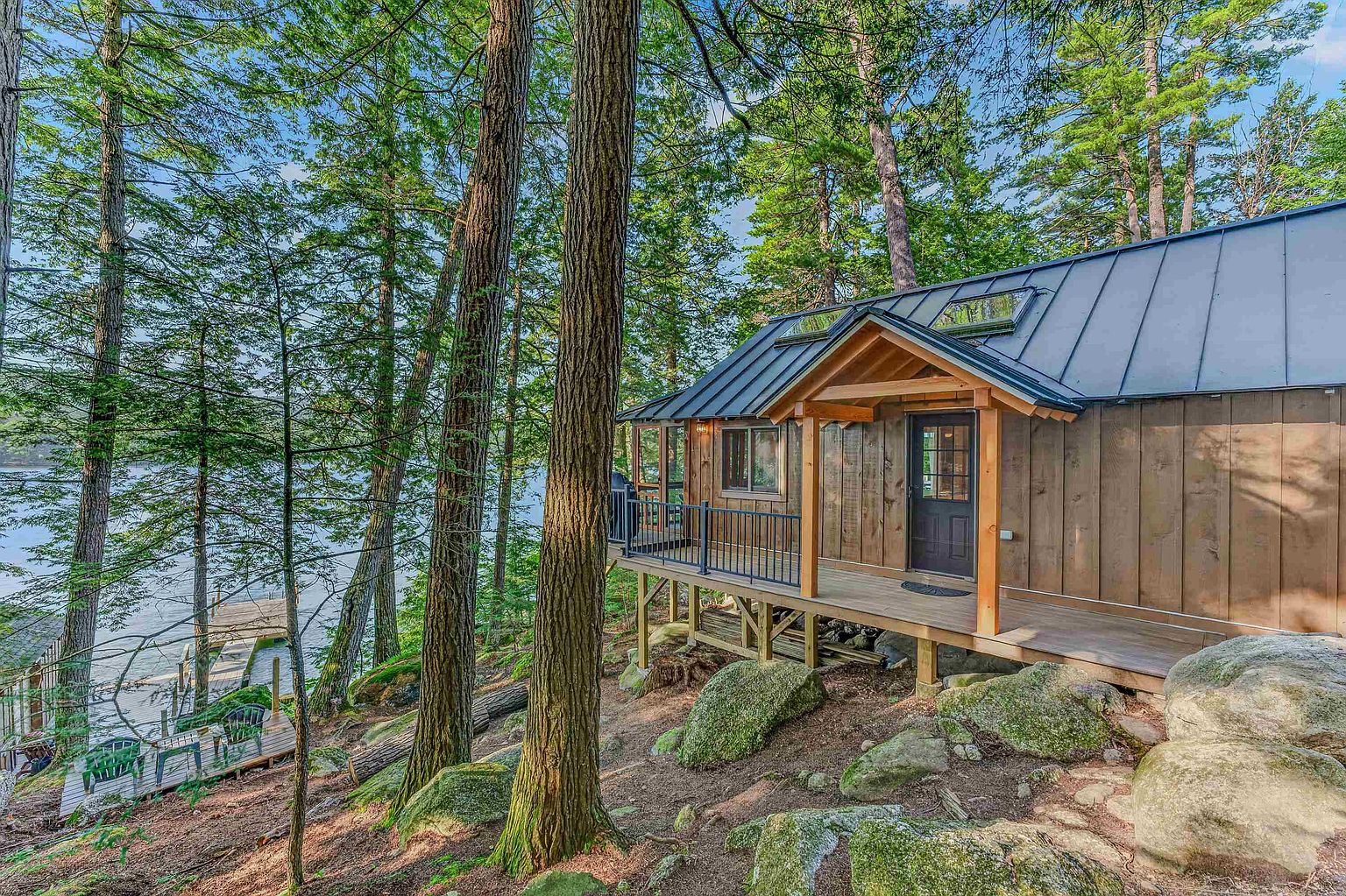 This rustic cabin is nestled within a dense, wooded forest, featuring vertical wood siding and a dark metal roof. A spacious wooden deck with a black metal railing wraps around the side, providing a scenic vantage point overlooking the adjacent lake and a private dock. The natural, rugged landscape is highlighted by large mossy boulders and mature pine trees, creating a serene and secluded atmosphere.