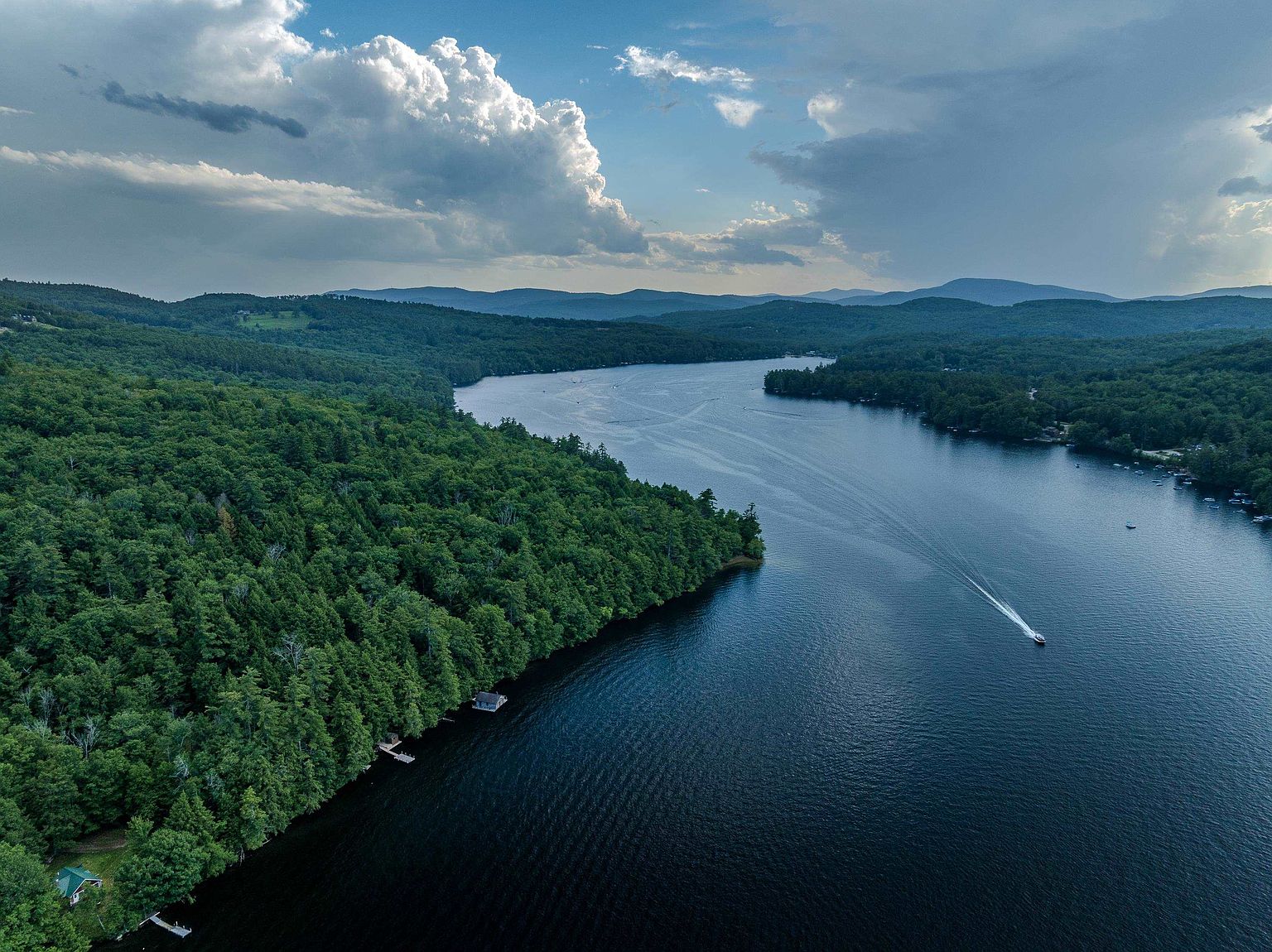 This stunning aerial perspective captures a vast, serene lake surrounded by dense, lush green forests and rolling mountains in the distance. A single motorboat leaves a gentle wake across the deep blue water, emphasizing the peaceful and expansive nature of the landscape. The scene is framed by a dramatic, cloud-filled sky, highlighting the property's prime location for waterfront recreation and natural beauty.