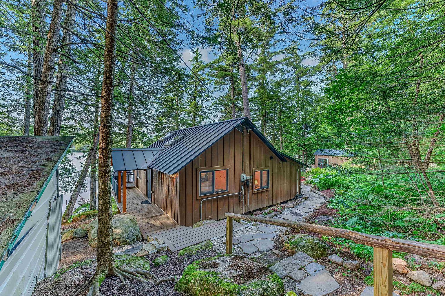This side view of a rustic cabin showcases vertical wood siding and a dark metal roof nestled within a lush, wooded landscape. A wooden walkway leads toward the structure, complemented by a stone path that winds through the mossy, natural terrain. The setting offers a serene, secluded atmosphere with mature trees providing a canopy over the home.