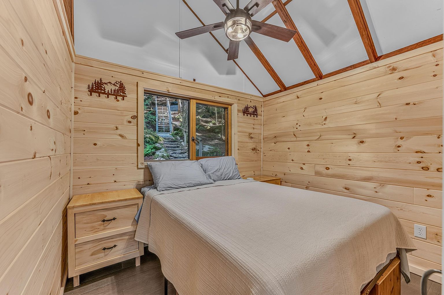 This cozy bedroom features light wood paneled walls and a vaulted ceiling with a central ceiling fan, creating a rustic cabin aesthetic. A bed with neutral bedding is positioned against the wall, flanked by a matching light wood nightstand, while a window provides a view of the wooded exterior. The room is compact and warm, emphasizing a natural, retreat-like atmosphere.
