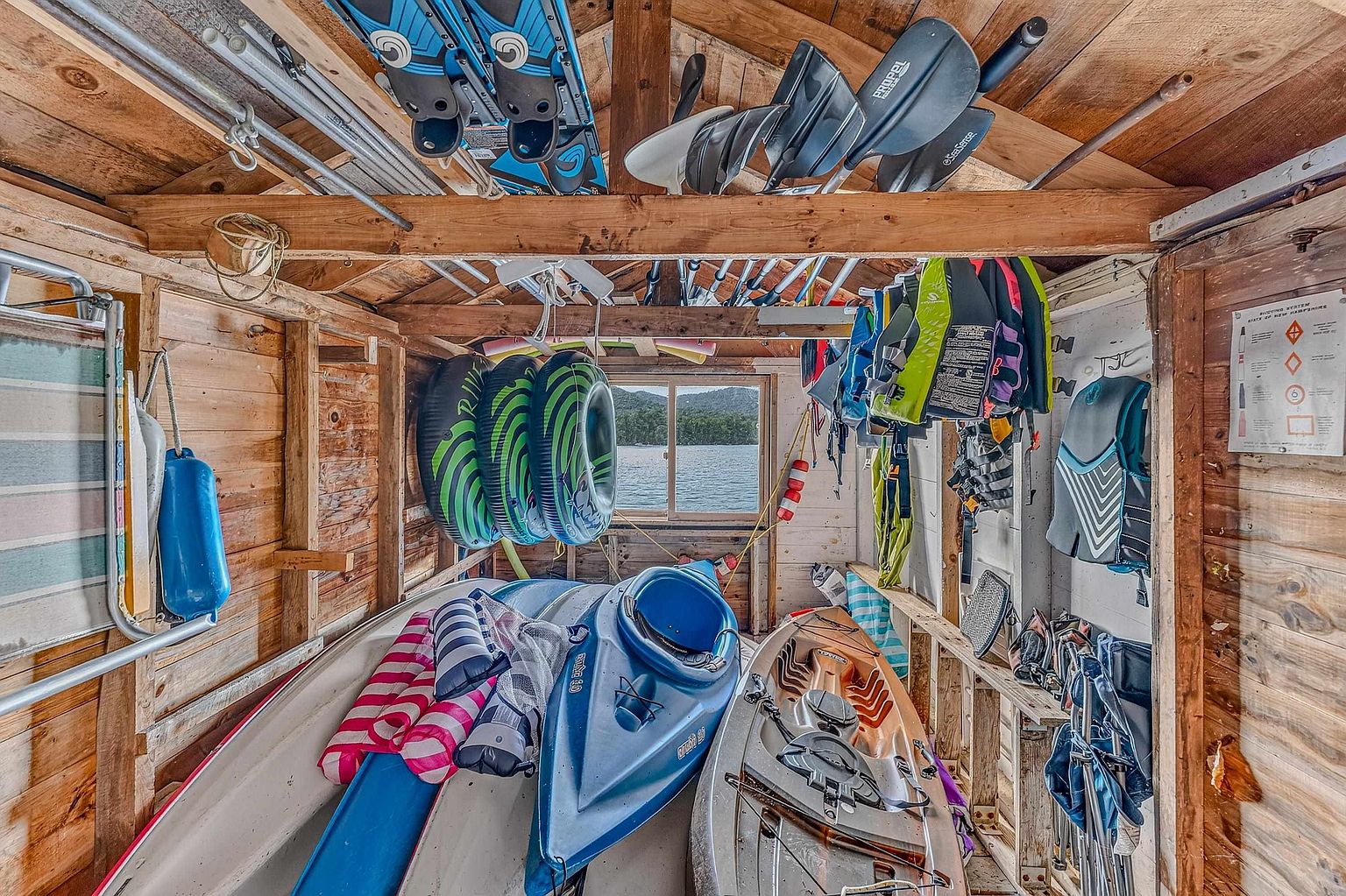 This rustic, wood-paneled storage shed is packed with recreational water equipment, including kayaks, paddles, life vests, and inflatable tubes. The space features exposed wooden rafters and walls, with a central window providing a direct view of the lake outside. The perspective is from the interior looking out, capturing a functional and organized storage area for waterfront activities.