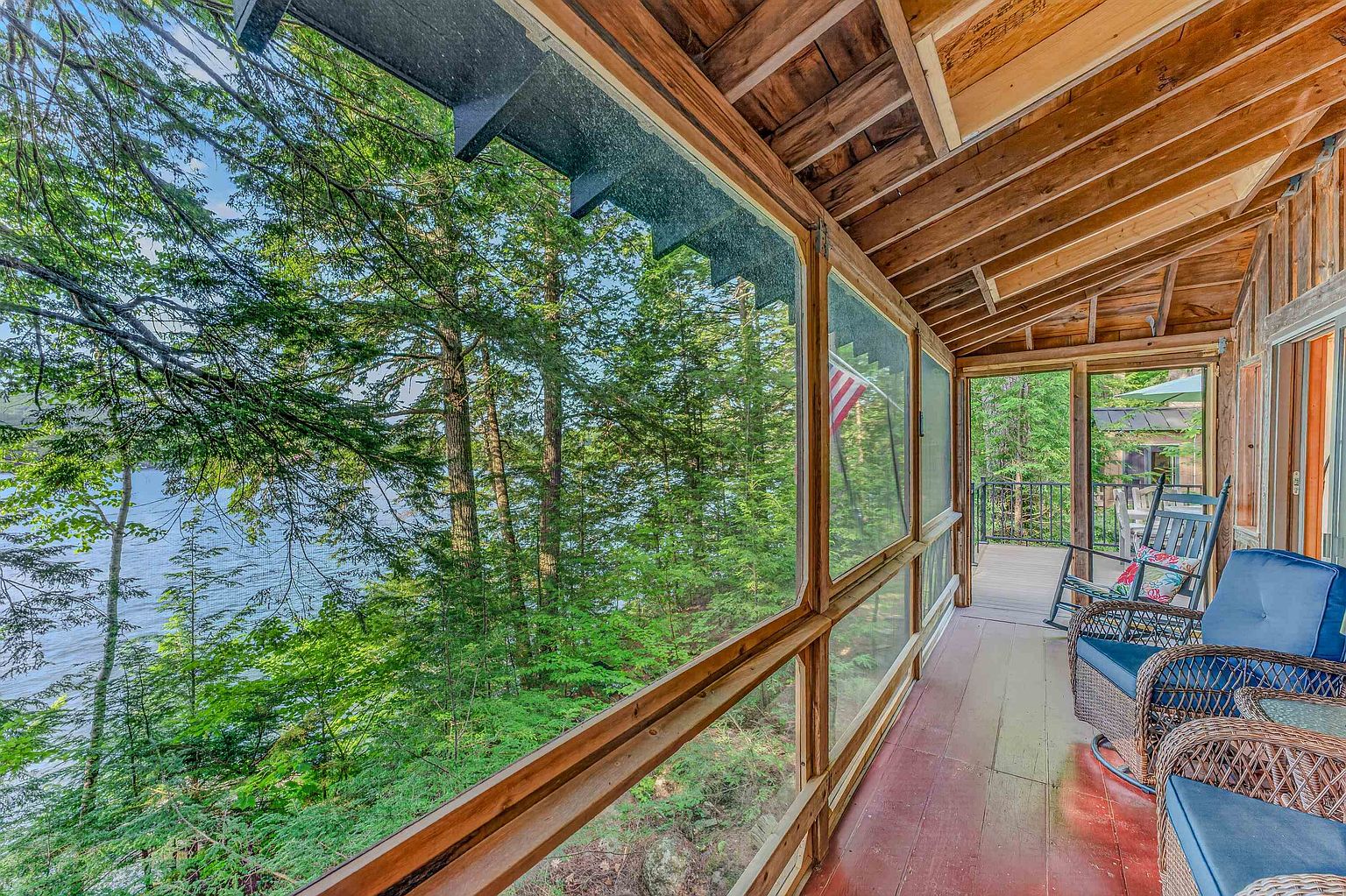 This screened-in porch offers a serene, elevated view of a lush forest and a nearby lake. The space features rustic wood framing, a painted floor, and comfortable wicker seating, creating a peaceful retreat that blends indoor comfort with the natural environment. The perspective looks out from the porch, emphasizing the seamless connection between the living area and the surrounding woodland scenery.