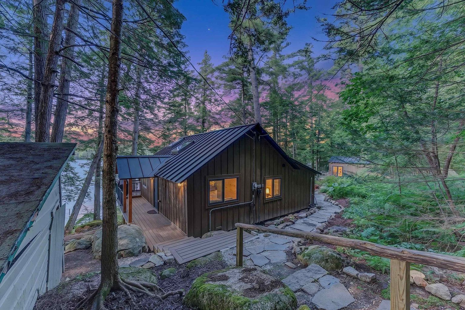 This side view captures a rustic, dark-toned cabin nestled within a lush, wooded landscape at twilight. The structure features a metal roof, a wooden deck extending toward the water, and warm interior lighting glowing through the windows. A stone pathway winds through the natural, rocky terrain, leading toward the home and a secondary structure visible in the background.