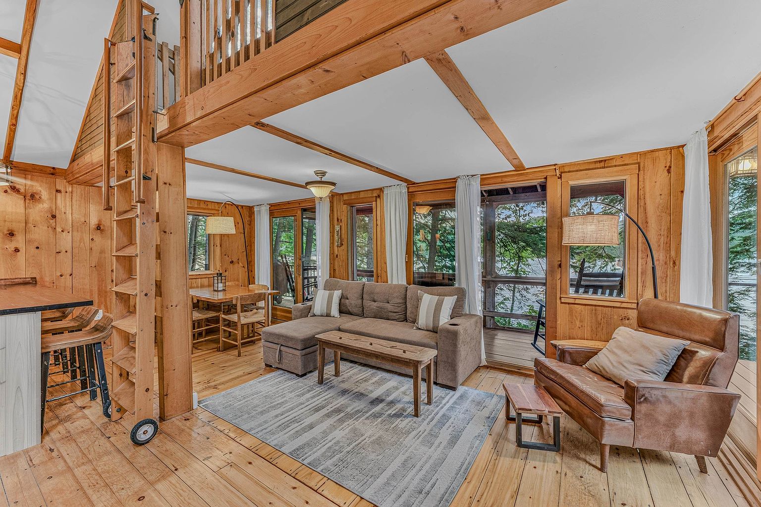 This cozy, rustic living room features warm wood-paneled walls and exposed ceiling beams, creating a cabin-like atmosphere. The space is furnished with a comfortable brown sectional sofa, a wooden coffee table, and a leather armchair, all arranged on a textured area rug. Large windows and a glass door provide ample natural light and views of the surrounding trees, while a wooden ladder leads to an upper loft area.