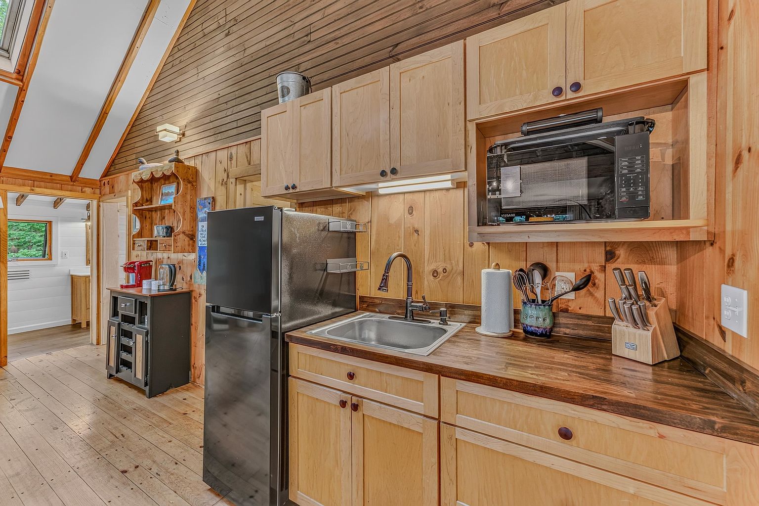 This rustic kitchen features light wood cabinetry and a dark wood countertop, creating a warm, cabin-like aesthetic. The space is equipped with a stainless steel sink, a black refrigerator, and a built-in microwave, all set against walls paneled in natural wood. A view into an adjacent room reveals a bright, white-walled space, while the vaulted ceiling adds a sense of openness to the compact cooking area.