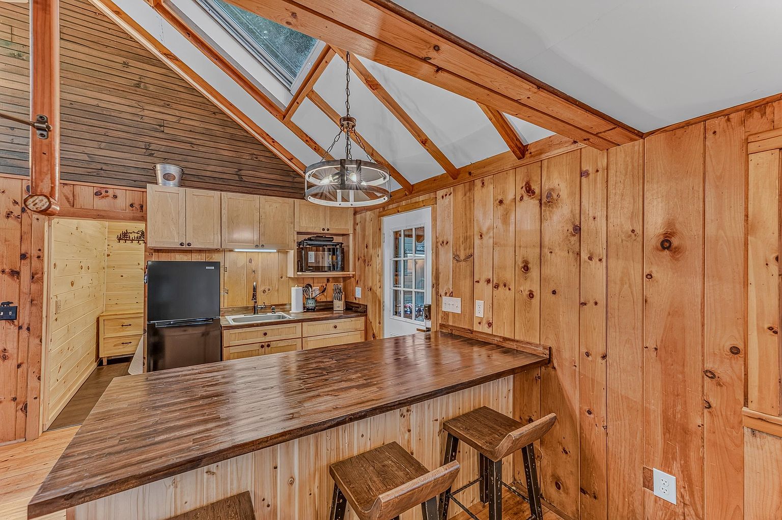 This rustic kitchen features light wood cabinetry, a dark wood breakfast bar, and a black refrigerator, creating a cozy cabin aesthetic. The space is highlighted by a vaulted ceiling with exposed wooden beams, a skylight, and a decorative chandelier hanging over the counter. A doorway leads to an adjacent room with matching wood-paneled walls, emphasizing the warm, natural wood theme throughout the home.