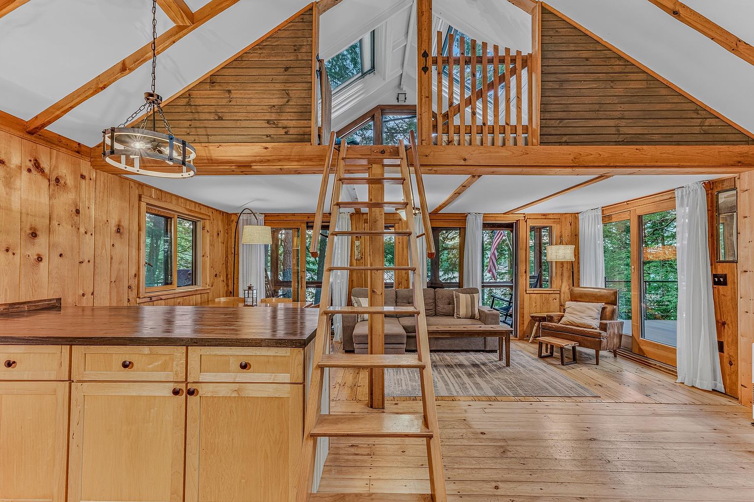 This rustic, open-concept living space features warm wood-paneled walls and a vaulted ceiling with exposed beams. A central wooden ladder leads to a loft area, while the main floor includes a cozy seating area with a sofa and leather armchair, all oriented toward large windows that provide a view of the surrounding trees. The foreground showcases a kitchen island with a dark countertop, creating a seamless transition between the culinary and relaxation zones.