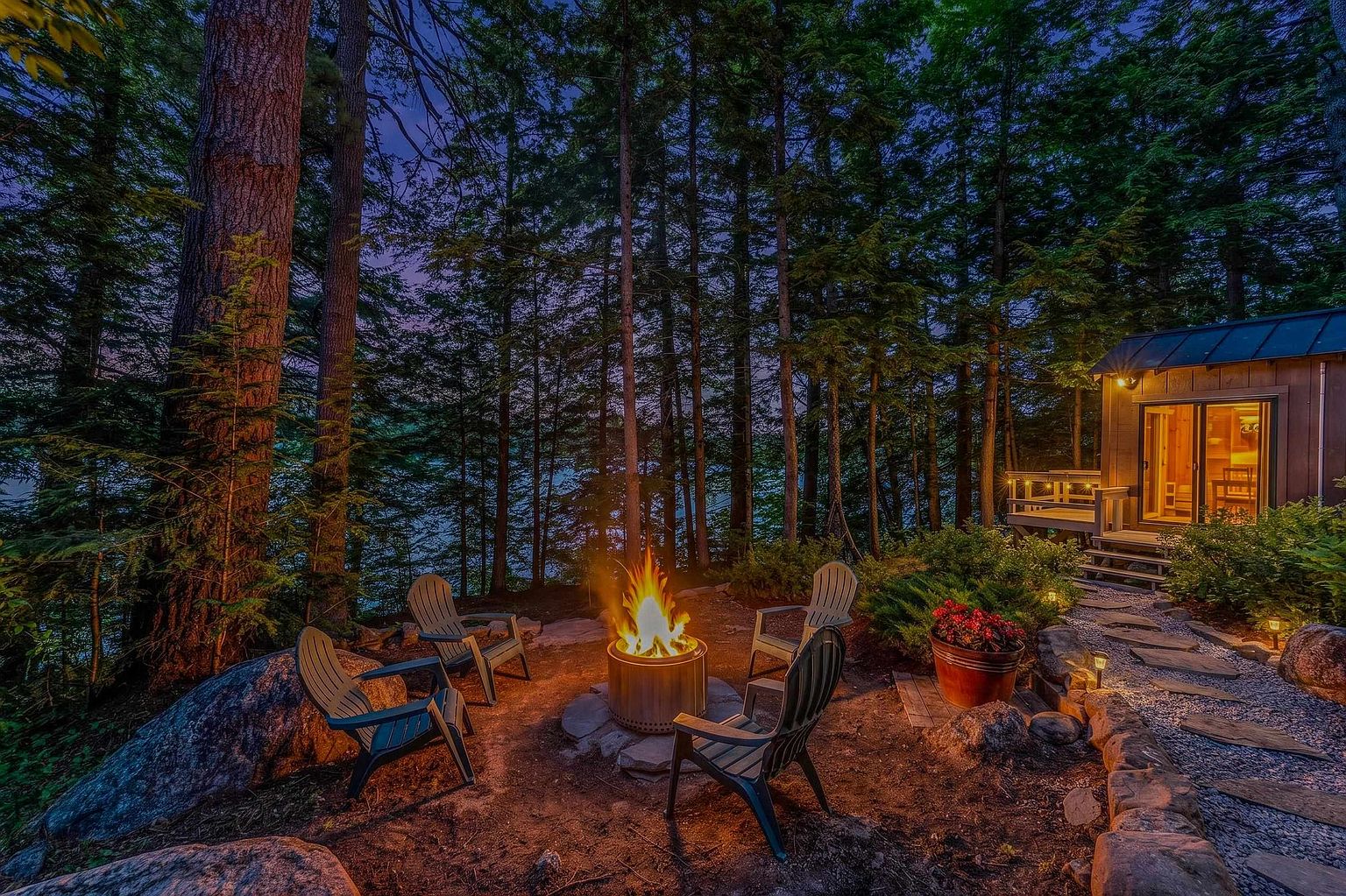 This serene outdoor space features a cozy fire pit area surrounded by four Adirondack chairs, set against a backdrop of dense, mature forest and a glimpse of a lake. A stone-paved path leads from the fire pit to a small, modern cabin with warm interior lighting visible through its glass doors. The scene is captured at twilight, creating an inviting and tranquil atmosphere perfect for evening relaxation.