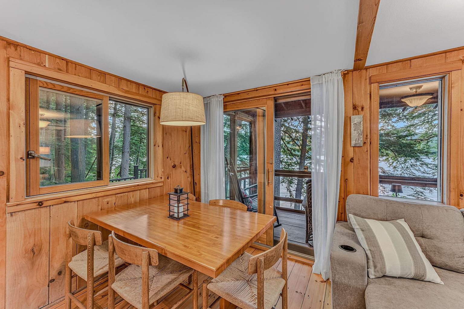 This cozy dining area features warm wood-paneled walls and a matching wooden table surrounded by four chairs with woven seats. A large pendant light hangs above the table, while expansive windows and a sliding glass door provide abundant natural light and scenic views of the surrounding forest and lake. The space is adjacent to a comfortable sofa, creating an inviting, rustic atmosphere perfect for relaxed meals.
