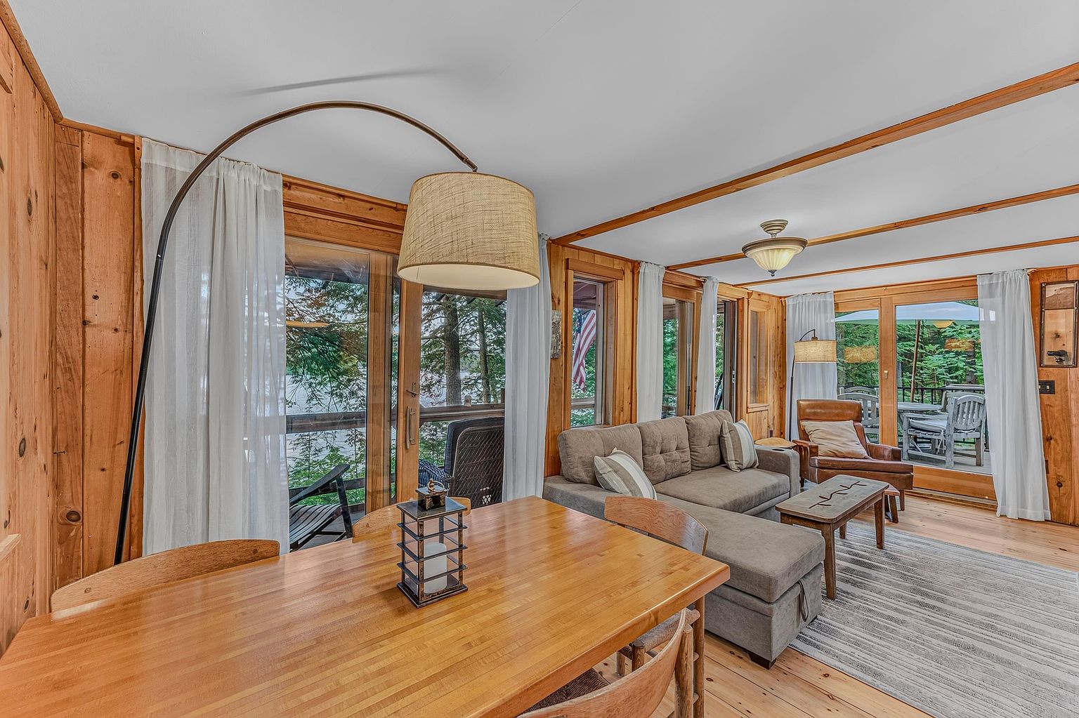 This cozy, rustic-style living and dining area features warm wood-paneled walls and ceilings, creating a cabin-like atmosphere. The space is anchored by a comfortable grey sectional sofa and a leather armchair, with large glass doors that provide expansive views of the surrounding trees and lake. A wooden dining table sits in the foreground, illuminated by a large, arched floor lamp, while the open-concept layout flows seamlessly toward the outdoor deck.