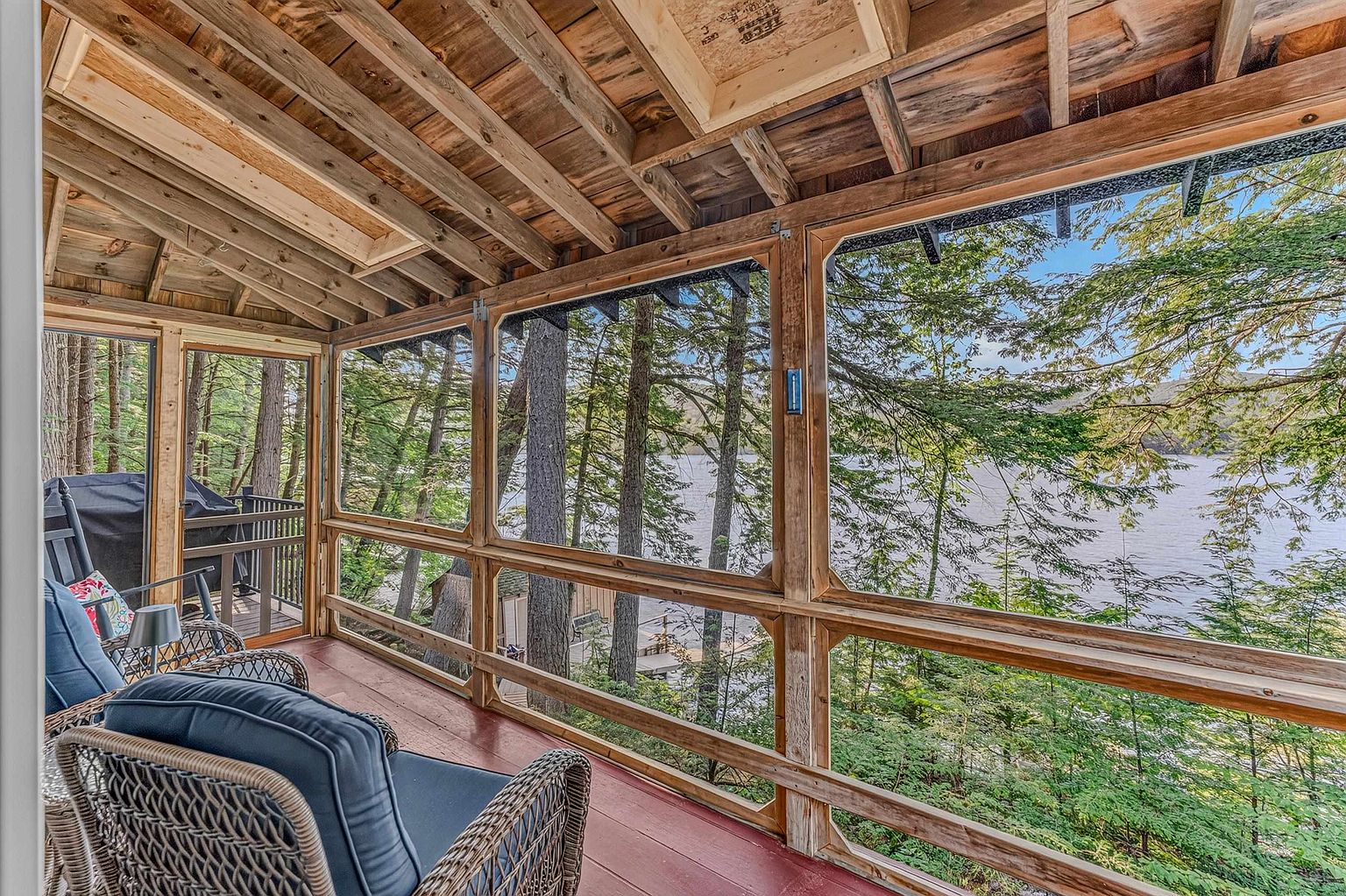 This screened-in porch offers a serene, rustic retreat with a clear view of the surrounding forest and lake. The space features exposed wooden rafters, a painted deck floor, and comfortable wicker seating, creating an inviting atmosphere for relaxation. Large screen panels provide an immersive connection to the outdoors while maintaining a sheltered, comfortable environment.