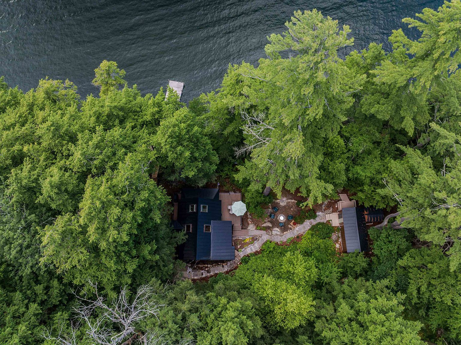 This high-angle aerial view captures a secluded cabin nestled within a dense, lush forest along a waterfront. The property features a dark-roofed main structure with an adjacent deck, a stone pathway leading to a secondary building, and a small private dock extending into the dark water. The perspective emphasizes the serene, wooded setting and the integration of the architecture into the natural landscape.