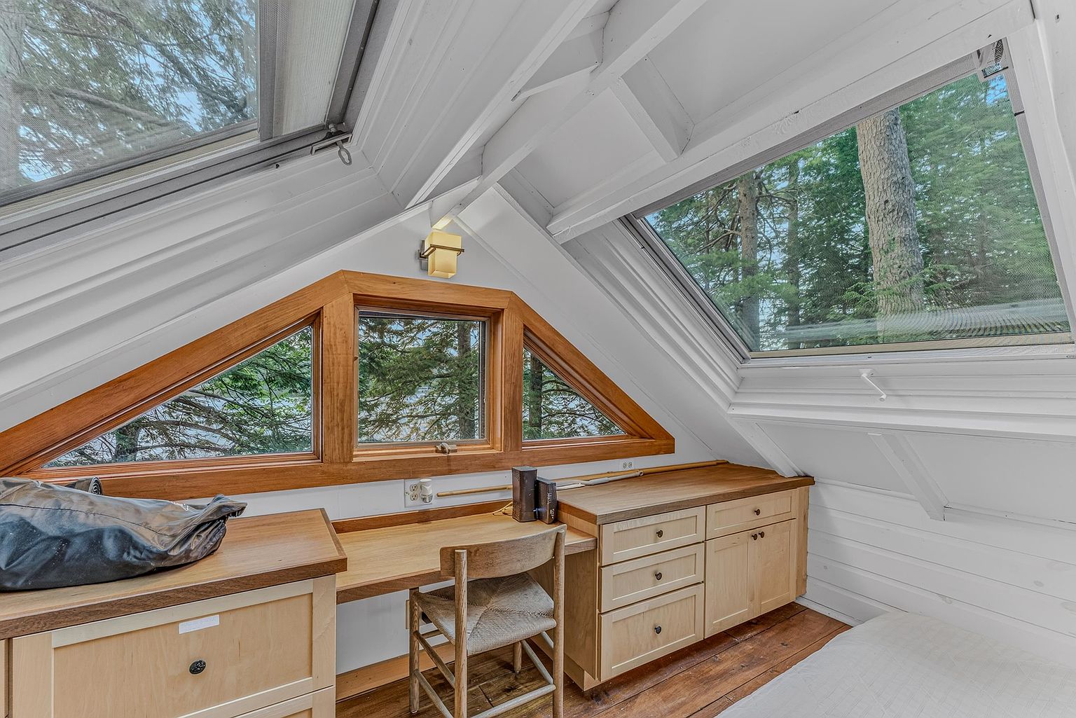 This cozy attic office space features a built-in light wood desk with integrated drawers and a matching chair, nestled under a sloped ceiling. A unique, multi-paneled triangular window provides a scenic view of the surrounding trees, while a large skylight above adds natural brightness to the room. The warm wood flooring and clean white walls create a peaceful, rustic atmosphere perfect for a quiet workspace.