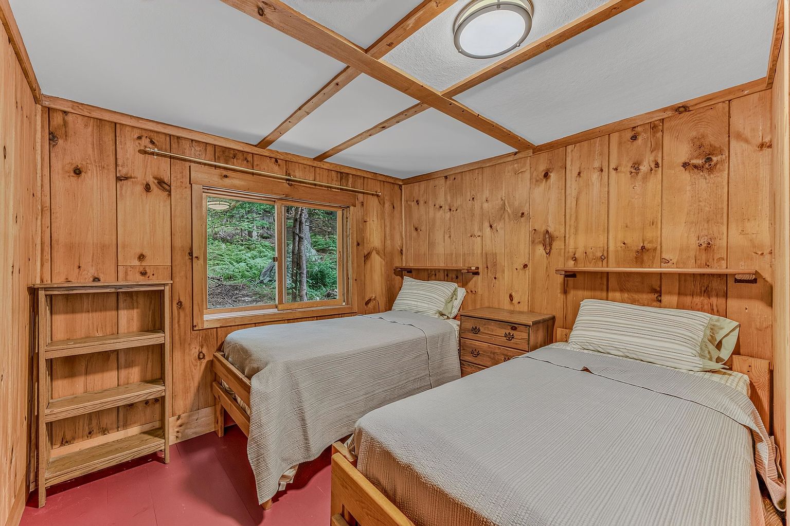 This cozy guest bedroom features rustic wood-paneled walls and a matching wood-beamed ceiling, creating a warm, cabin-like atmosphere. The room is furnished with two twin beds, a central wooden nightstand, and a tall wooden bookshelf, all set against a painted floor. A window provides a view of the surrounding forest, bringing natural light into the space.