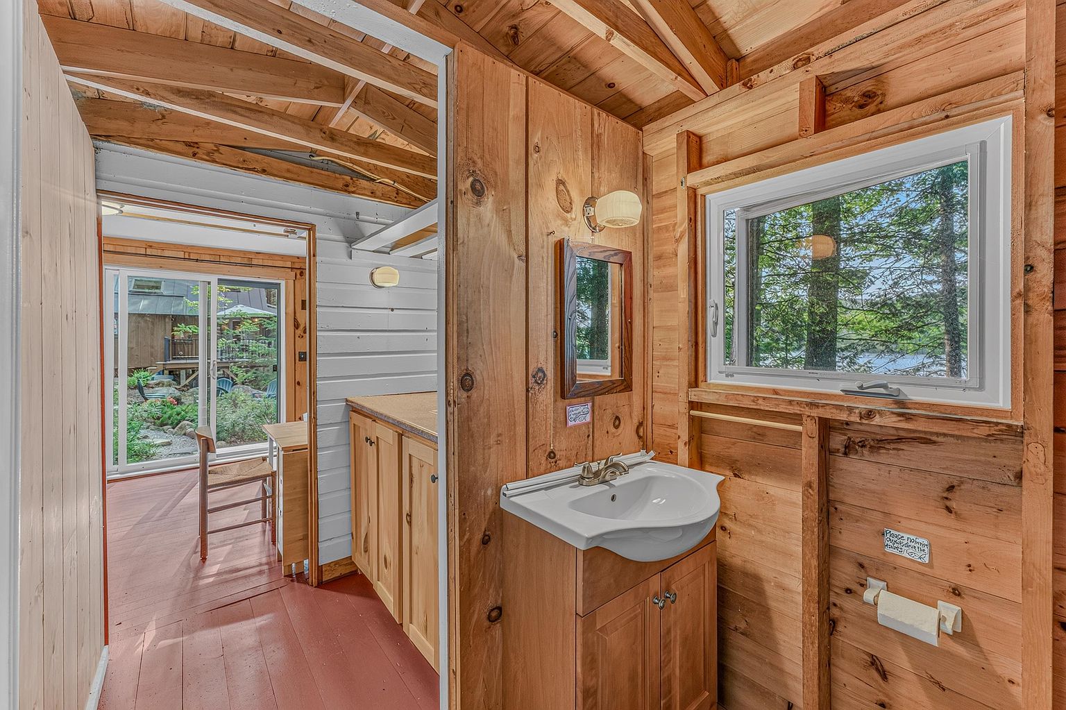 This rustic bathroom features warm wood-paneled walls and a matching vanity with a white sink. A window above the vanity provides natural light and a view of the surrounding trees, while a doorway leads to an adjacent room with a wooden floor and sliding glass doors. The space has a cozy, cabin-like aesthetic with exposed wooden ceiling beams.