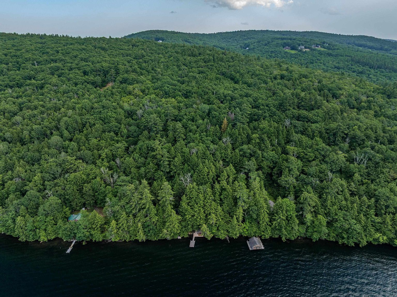 This aerial perspective captures a secluded waterfront property nestled within a dense, lush forest. The scene features a small cabin partially hidden by trees, a private wooden dock extending into the dark water, and a small boathouse structure along the shoreline. The rolling, tree-covered hills in the background provide a serene and private natural setting.