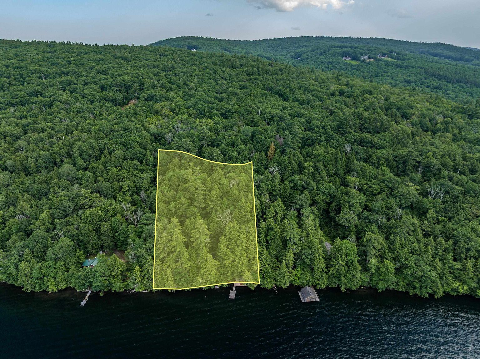 This aerial perspective captures a heavily wooded waterfront lot highlighted by a yellow boundary outline. The property features direct access to the lake, including a private dock, and is surrounded by dense, mature forest that extends up the hillside. The scene conveys a sense of seclusion and natural beauty, ideal for a private retreat.