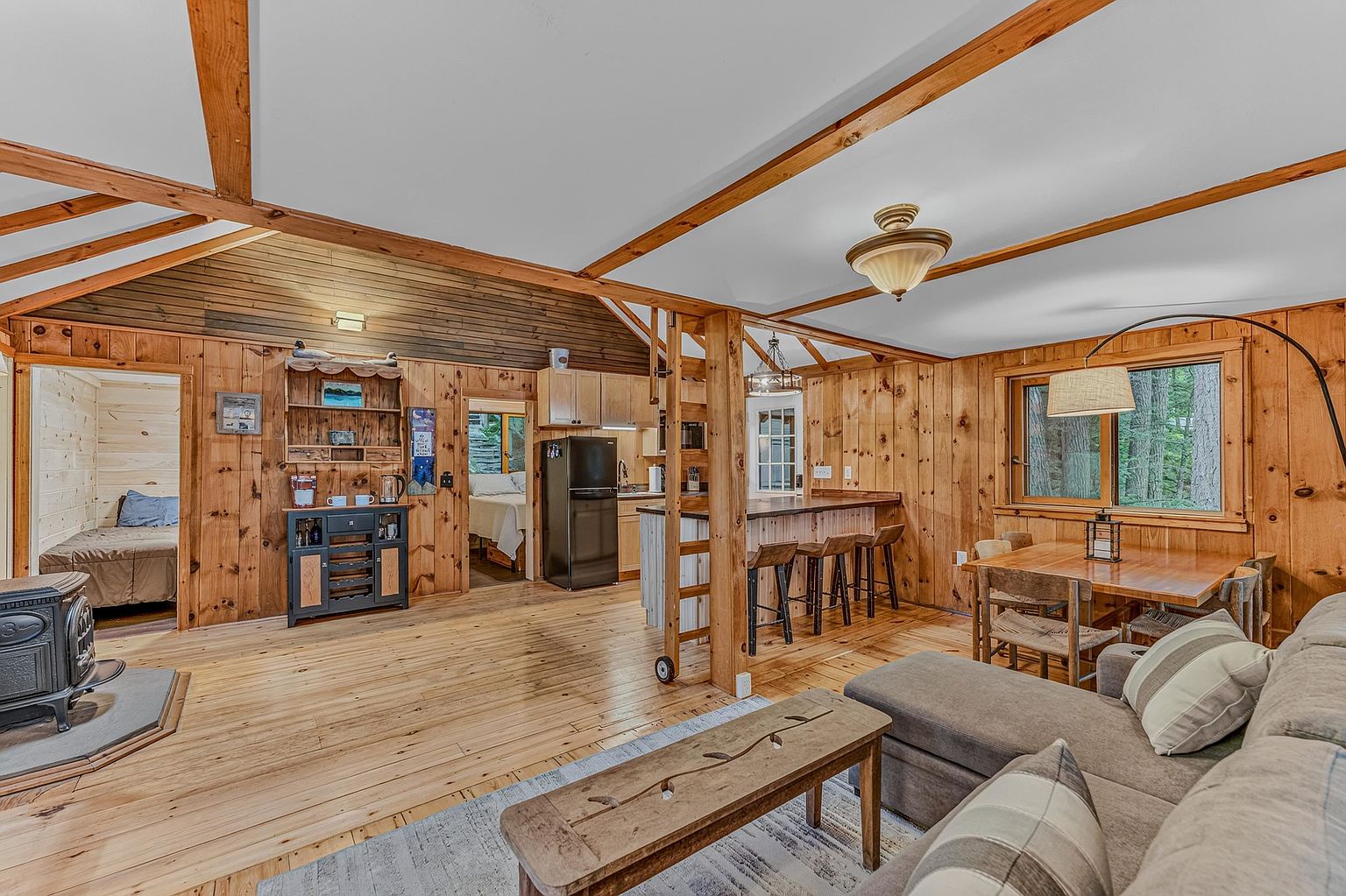 This open-concept living space features warm wood-paneled walls and exposed ceiling beams, creating a cozy, rustic cabin atmosphere. The room includes a comfortable sectional sofa, a wooden coffee table, and a wood-burning stove on a stone hearth, while a breakfast bar connects the living area to the kitchen. The perspective captures the flow between the main living area, the kitchen, and two visible bedroom doorways, highlighting the home's inviting and functional layout.