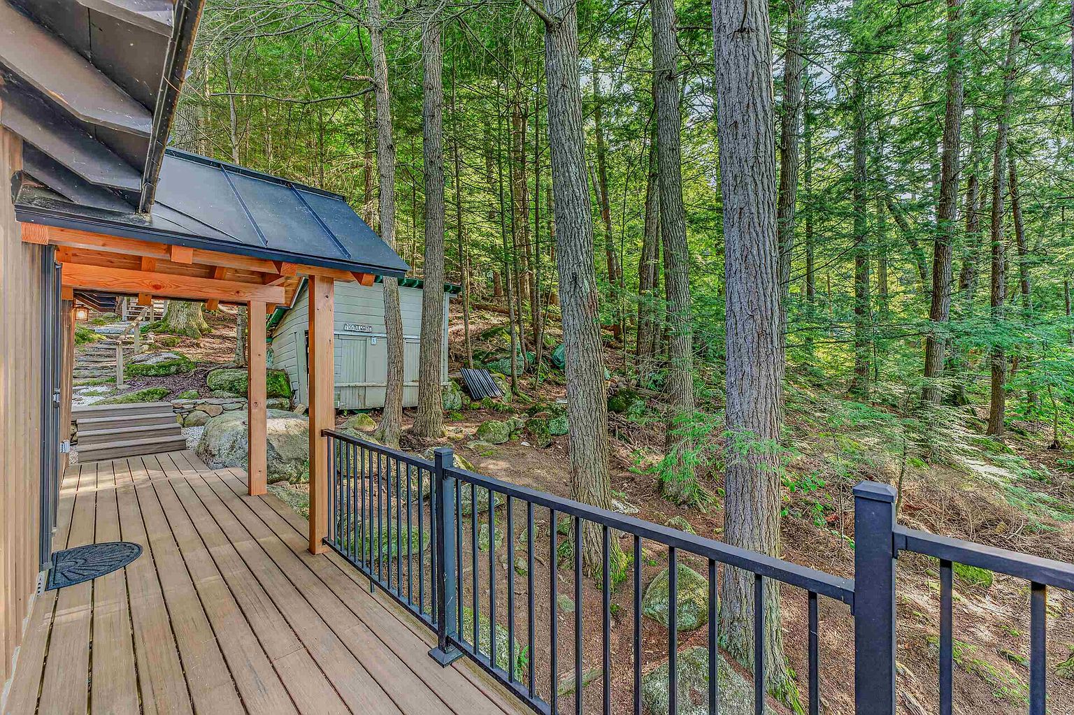 This image captures a wooden deck attached to a rustic home, featuring a black metal railing that overlooks a dense, wooded forest. The perspective is from the deck looking out toward a small, light-colored shed nestled among large boulders and mature trees. The scene conveys a peaceful, secluded atmosphere, highlighting the home's integration with the natural landscape.