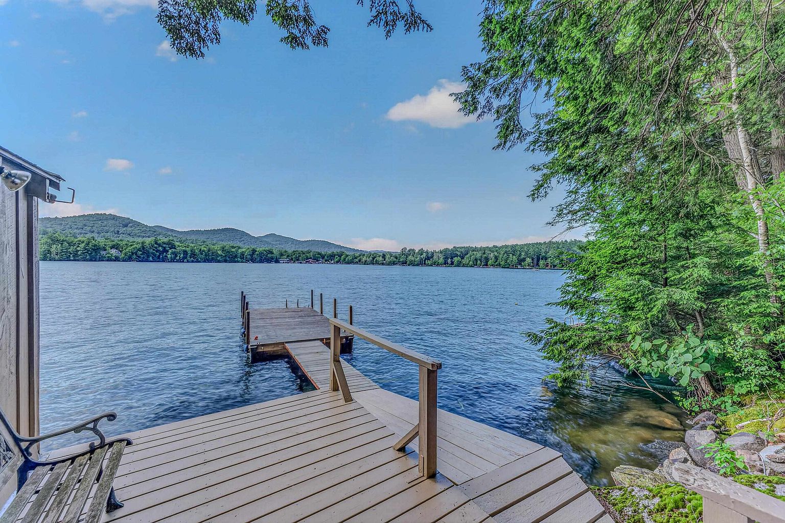 This image captures a serene lakeside deck featuring a wooden platform that extends into a private dock. The perspective looks out over calm, deep blue water toward a distant, tree-covered mountain range under a bright, clear sky. Lush green trees frame the right side of the deck, creating a peaceful and secluded atmosphere perfect for waterfront relaxation.