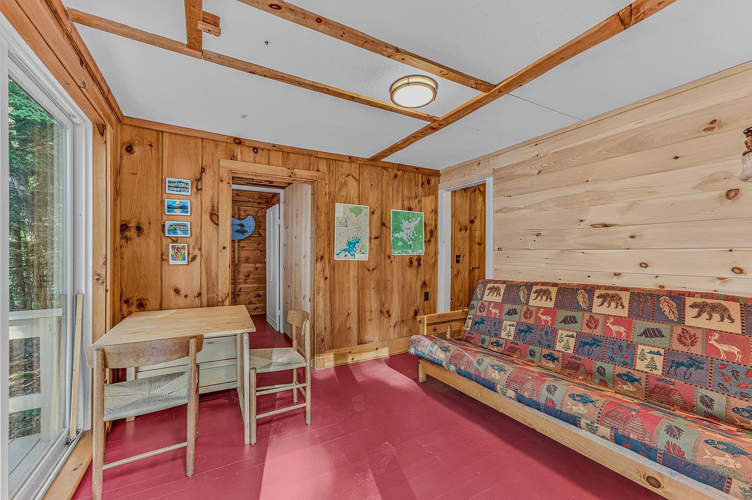 This rustic interior space features warm wood-paneled walls and a vibrant red painted floor, creating a cozy, cabin-like atmosphere. The room is furnished with a futon featuring a patterned wildlife-themed fabric and a simple wooden table with two chairs. Large sliding glass doors on the left provide ample natural light and a view of the surrounding trees, while maps and framed art decorate the walls.