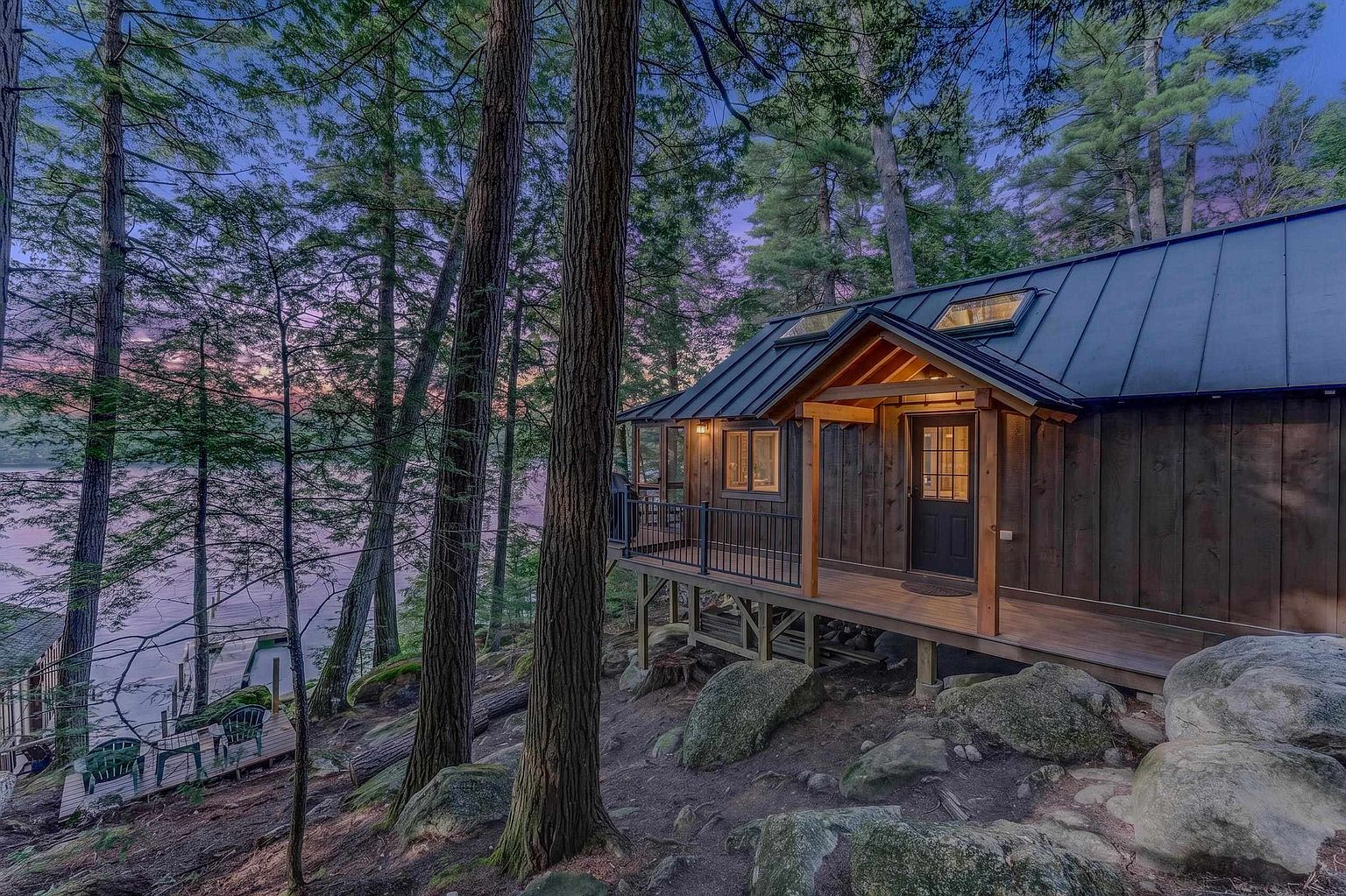 This rustic cabin is nestled among tall pine trees on a rocky, wooded slope overlooking a serene lake at dusk. The structure features dark vertical wood siding, a metal roof with integrated skylights, and a covered wooden deck with a black metal railing. The twilight sky casts a soft purple and orange glow over the water, creating a peaceful and secluded atmosphere.