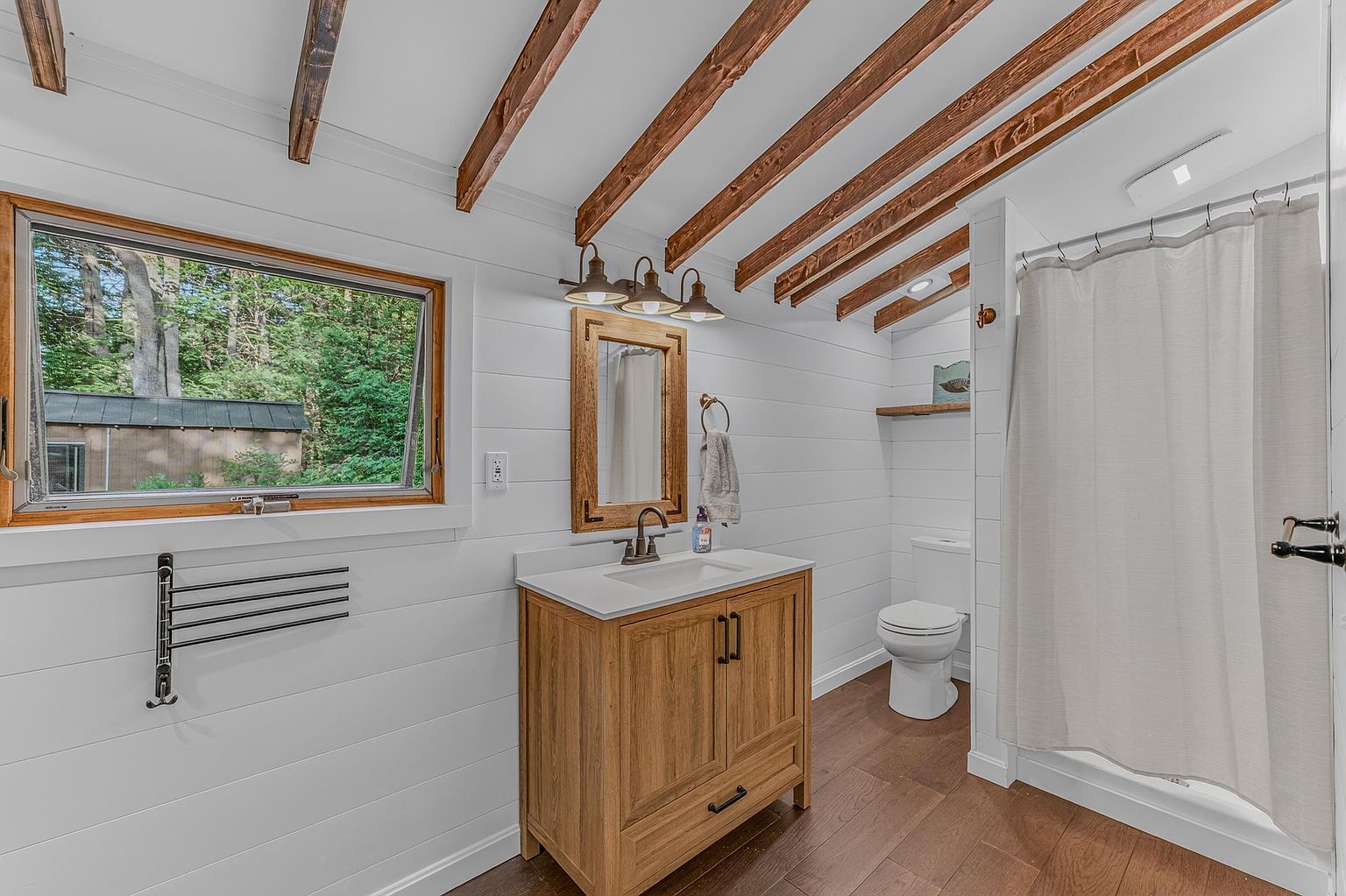 This bright, rustic-inspired bathroom features white shiplap walls and exposed wooden ceiling beams, creating a warm and inviting atmosphere. The space includes a light wood vanity with a white countertop, a framed mirror with matching lighting, and a walk-in shower with a neutral curtain. A large window provides natural light and a view of the surrounding greenery, while dark wood-toned flooring completes the cohesive design.