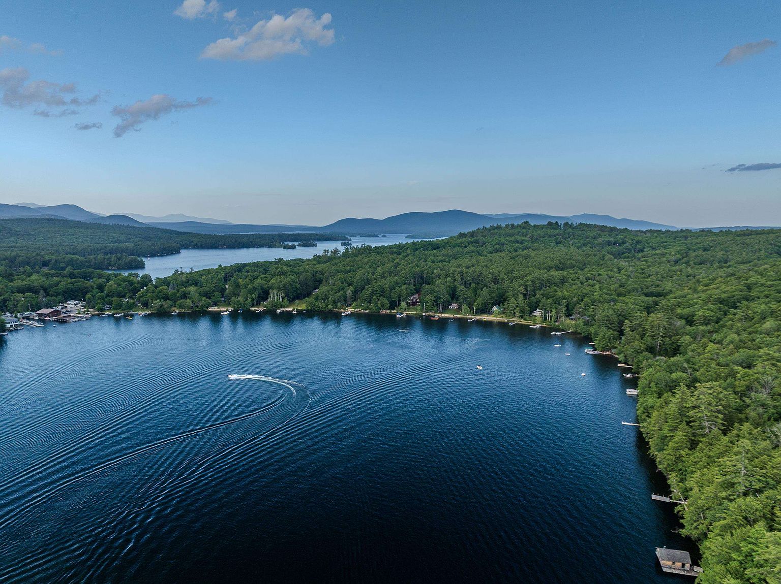 This high-angle aerial view captures a sprawling, deep blue lake surrounded by dense, lush green forests and distant rolling mountains under a clear sky. A small motorboat leaves a distinct wake across the water, while the shoreline is dotted with small docks and secluded waterfront properties nestled among the trees. The perspective provides a serene and expansive look at the natural landscape and the recreational potential of the waterfront location.