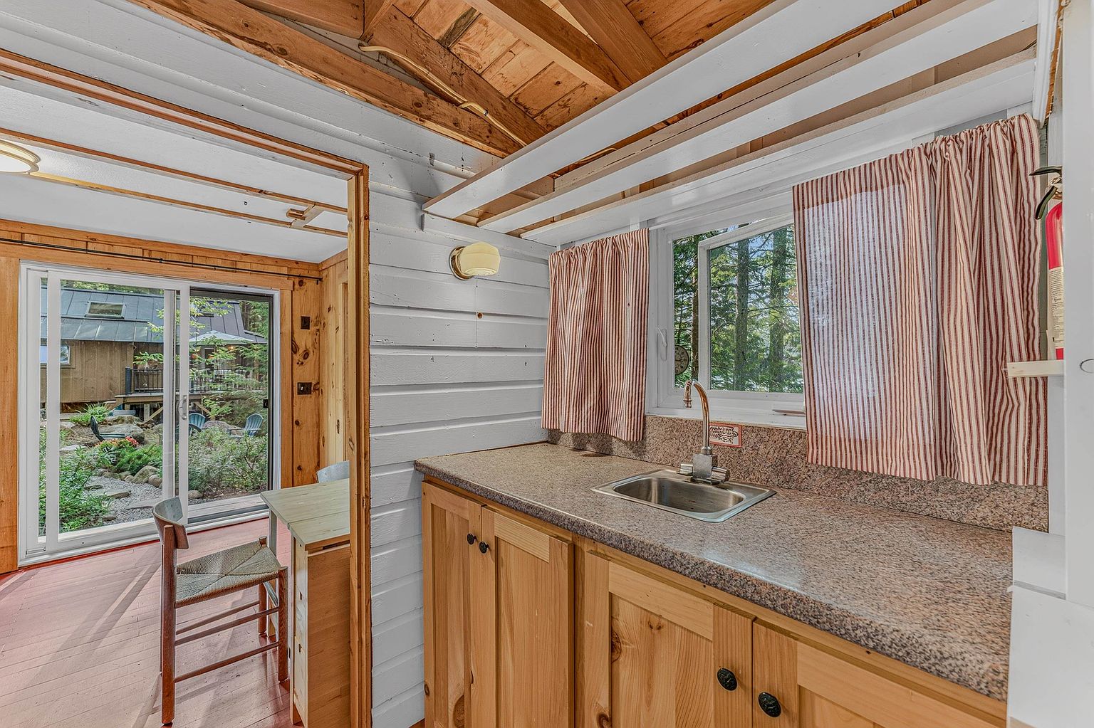 This cozy kitchen area features light wood cabinetry topped with a speckled stone countertop and a stainless steel sink. A window with striped curtains provides a view of the lush, wooded exterior, while a sliding glass door nearby opens to a deck, creating a seamless indoor-outdoor connection. The space is defined by rustic wood-paneled walls and a vaulted ceiling, offering a warm and inviting cabin-like atmosphere.
