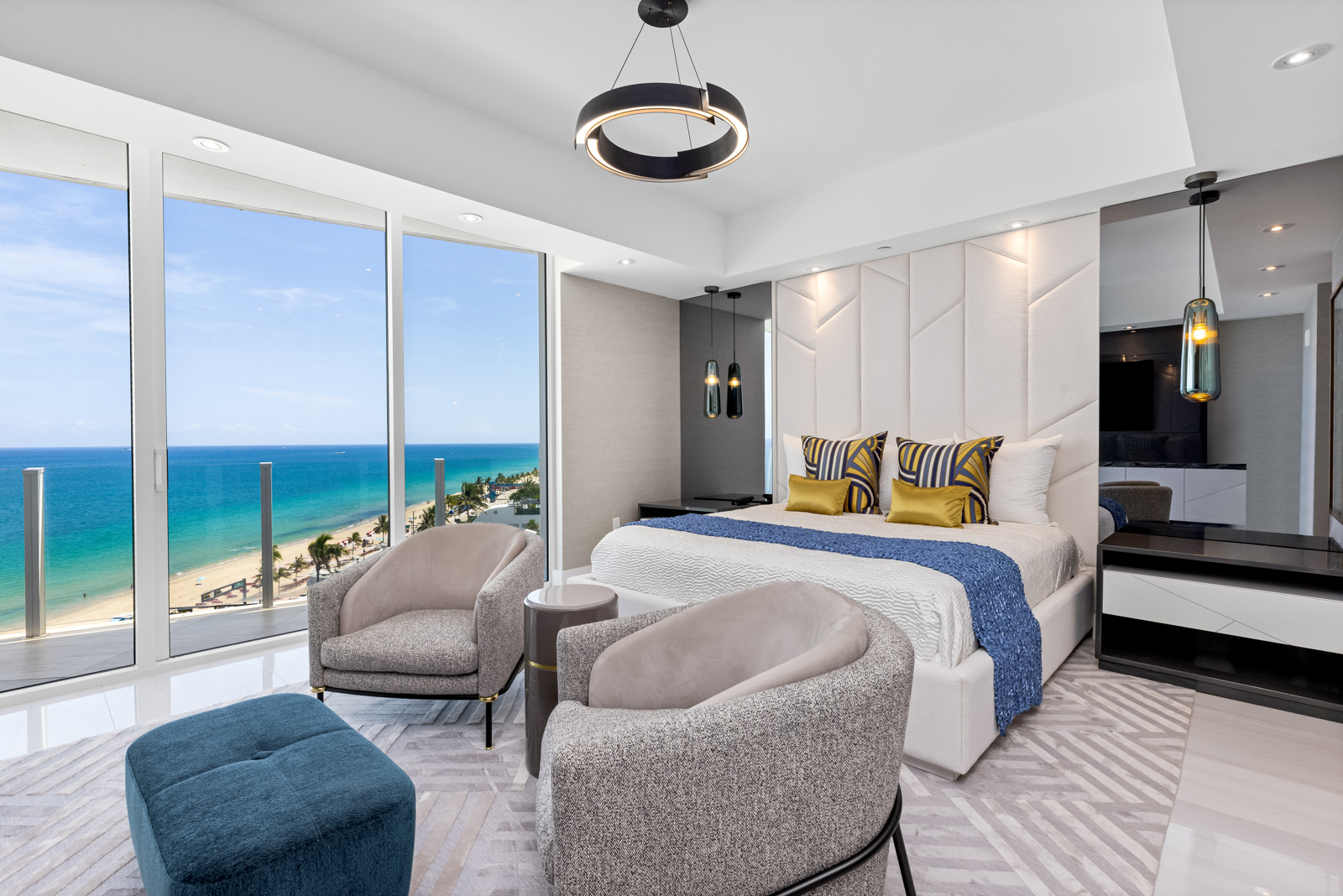 This is a luxurious primary bedroom featuring a king-size bed with a padded headboard and stylish throw pillows. The room has floor-to-ceiling windows offering ocean views, two modern armchairs, and a contemporary light fixture. The overall impression is one of elegance and comfort, perfect for a high-end real estate listing.