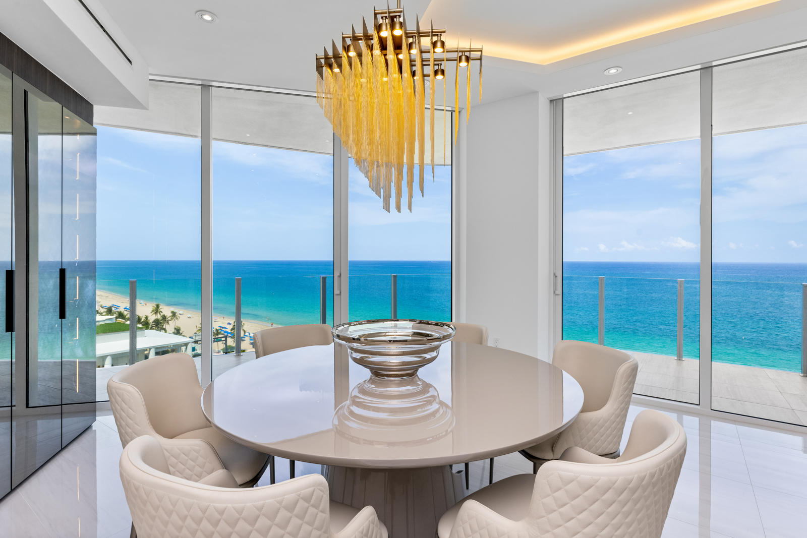 This is a bright and luxurious dining room featuring a round table with seating for six, surrounded by floor-to-ceiling windows offering stunning ocean views. A modern chandelier hangs above the table, adding a touch of elegance. The room's design emphasizes natural light and a seamless connection to the outdoor scenery, creating a sophisticated and inviting atmosphere.