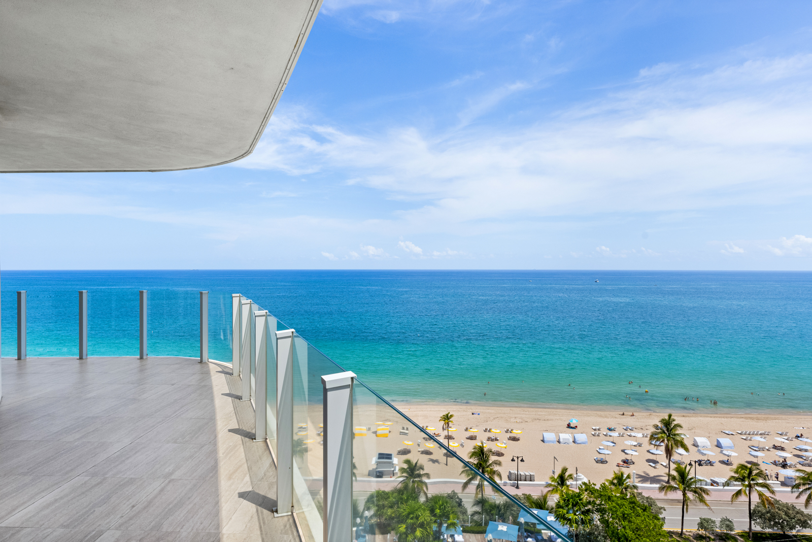This image showcases a luxurious balcony overlooking a beautiful beach and ocean. The balcony features a modern glass railing and a tiled floor, providing an unobstructed view of the turquoise water and sandy beach below. Palm trees and beach umbrellas dot the landscape, creating a serene and inviting atmosphere.