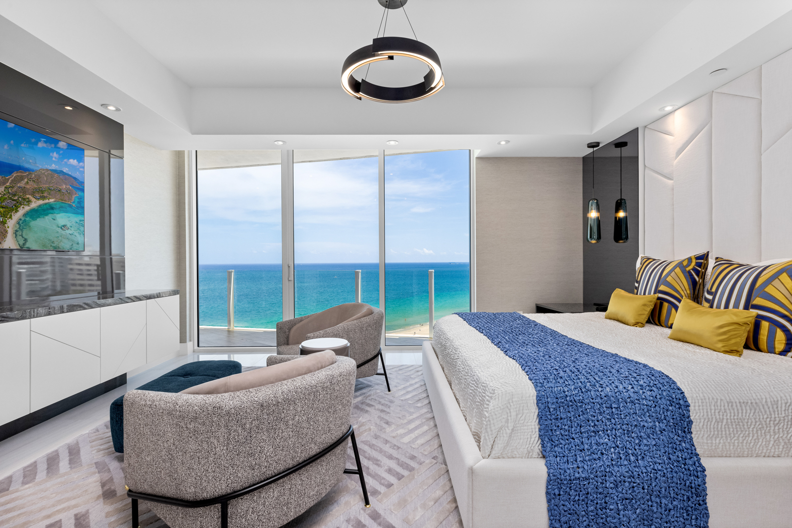 This is a luxurious primary bedroom with a stunning ocean view. The room features a large bed with a white upholstered headboard, stylish throw pillows, and a blue throw blanket. There are two modern armchairs and a small table near the floor-to-ceiling windows, and a sleek entertainment center with a large television. The overall impression is one of elegance and relaxation.