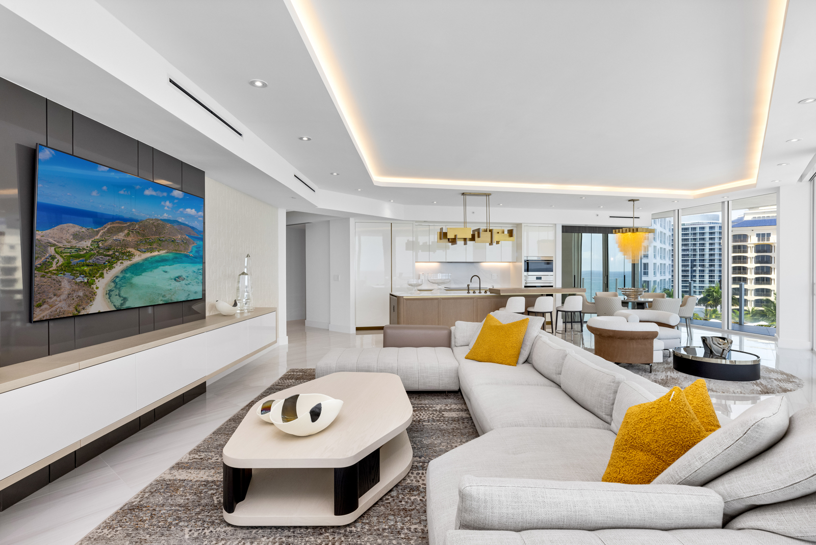 This is a bright and modern living room featuring a large sectional sofa with yellow accent pillows, a unique coffee table, and a flat-screen TV mounted on a stylish wall unit. The open-concept design seamlessly connects the living area to the kitchen and dining space, with floor-to-ceiling windows offering stunning city views. The overall impression is luxurious and contemporary.