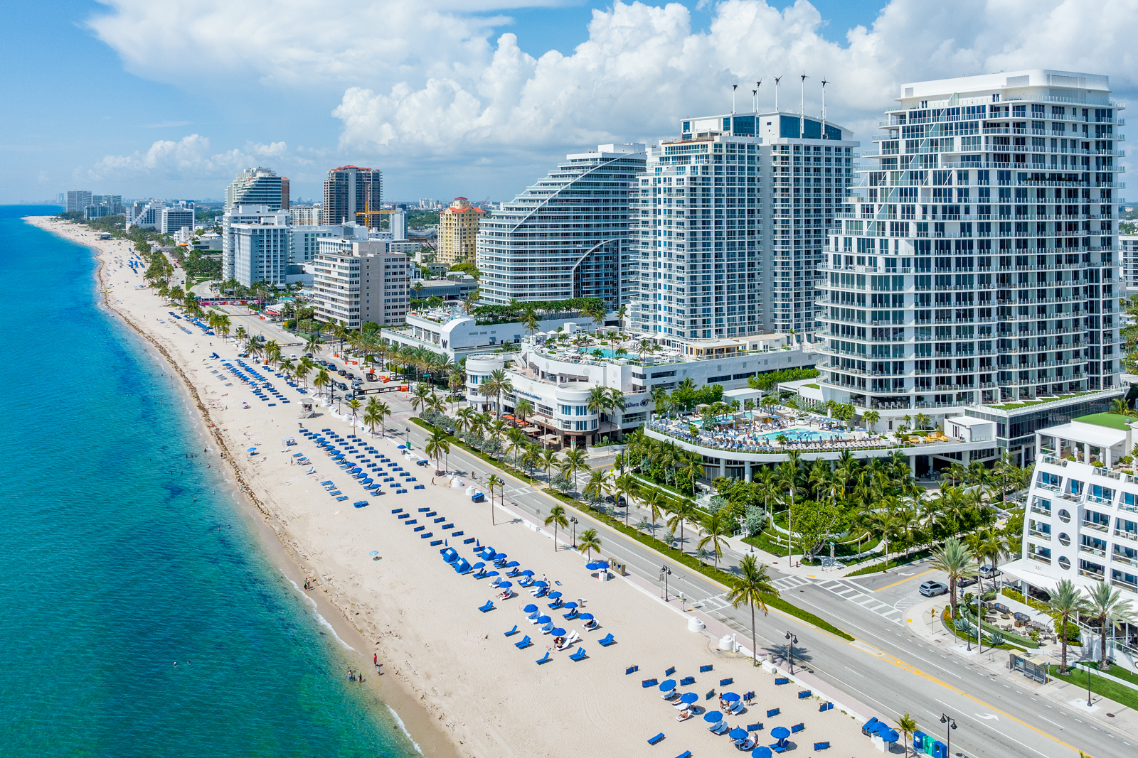 This aerial view showcases a stunning beachfront property with modern high-rise buildings and a pristine sandy beach. The turquoise ocean contrasts beautifully with the white buildings and blue beach chairs, creating a luxurious and inviting atmosphere. Palm trees line the street, adding to the tropical ambiance.