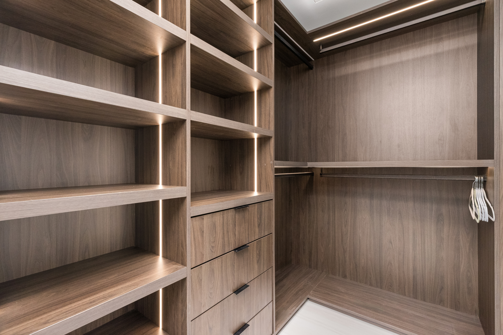 This image showcases a well-organized walk-in closet with custom shelving and drawers. The closet features a wood-grain finish, integrated lighting, and ample storage space for clothing and accessories. The design is modern and functional, offering a luxurious and efficient storage solution.